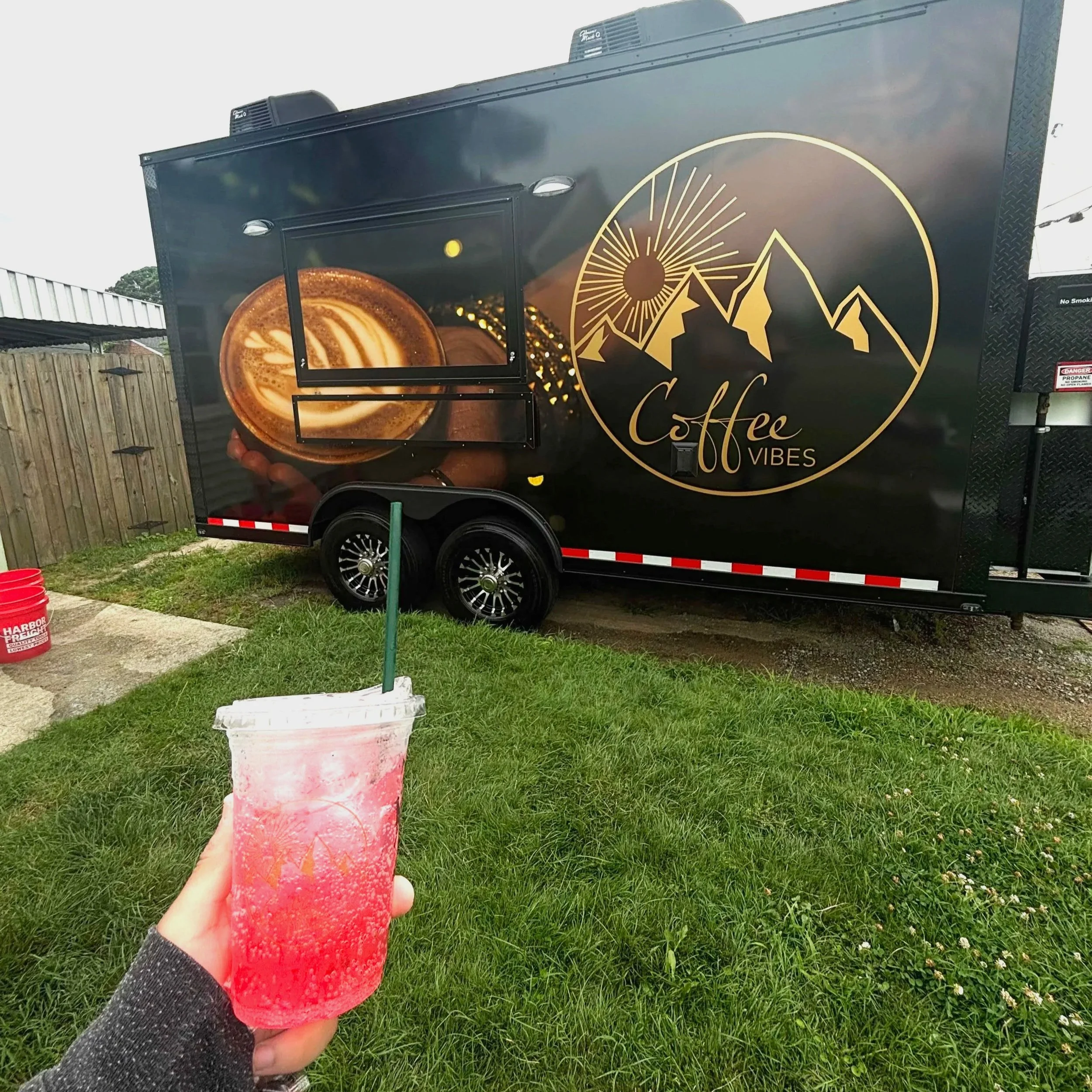 A person holding a pink iced beverage with a straw in front of a black coffee truck with gold mountain and sun design that reads "Coffee Vibes."