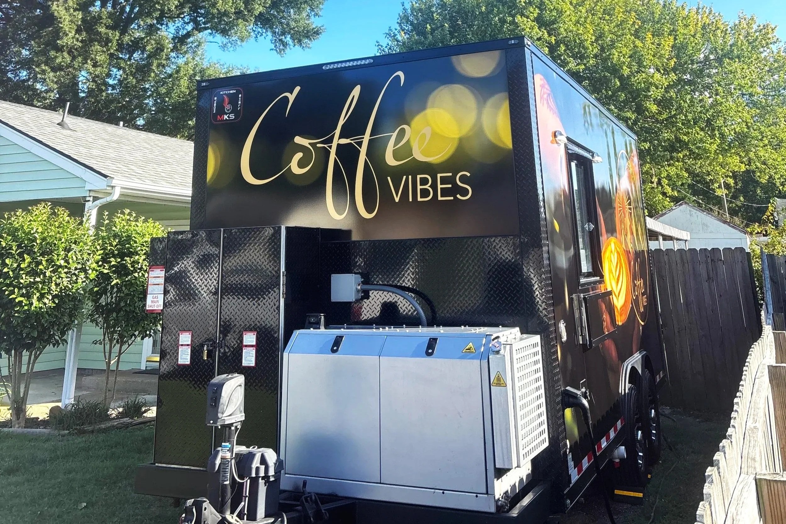 Black coffee truck with the words "Coffee Vibes" in cursive and capital letters, parked outdoors near a house surrounded by trees and a wooden fence.
