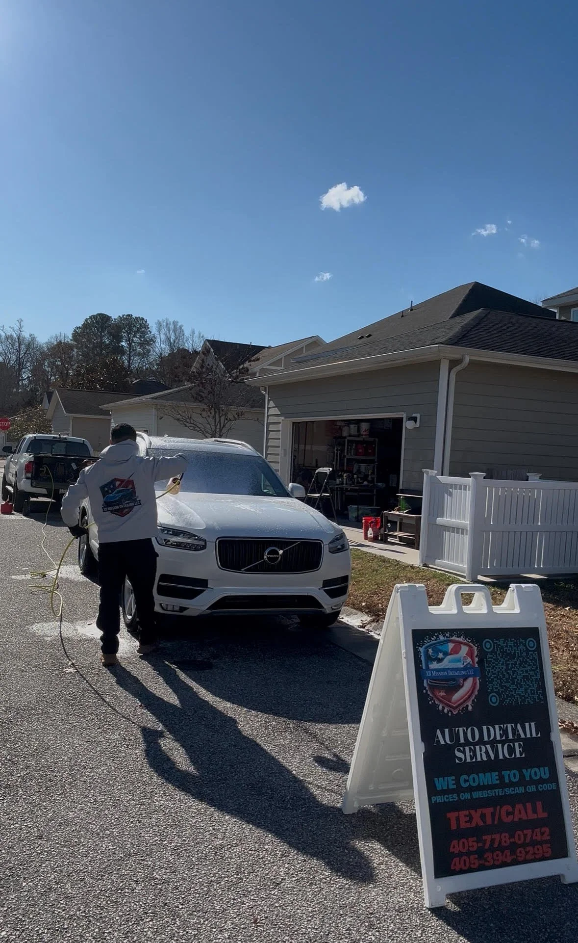 Person cleaning a white Volvo SUV outside a house, with an signboard for auto detail service that says 'We come to you,' phone numbers, and a QR code.