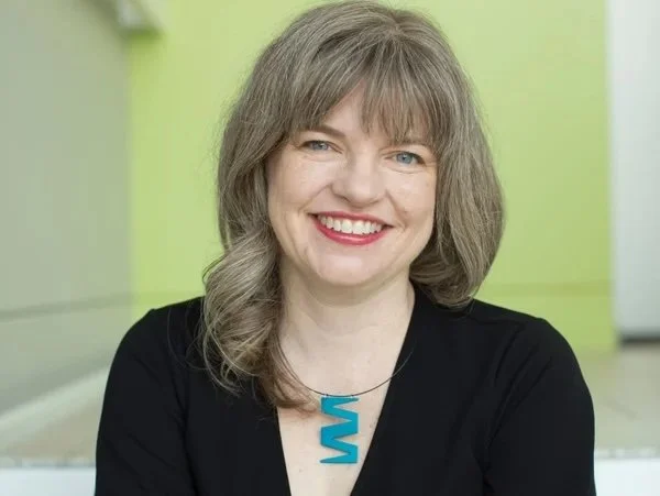 A woman with shoulder-length gray hair smiling, wearing a black top and a turquoise necklace, standing in a room with light green and yellow walls.