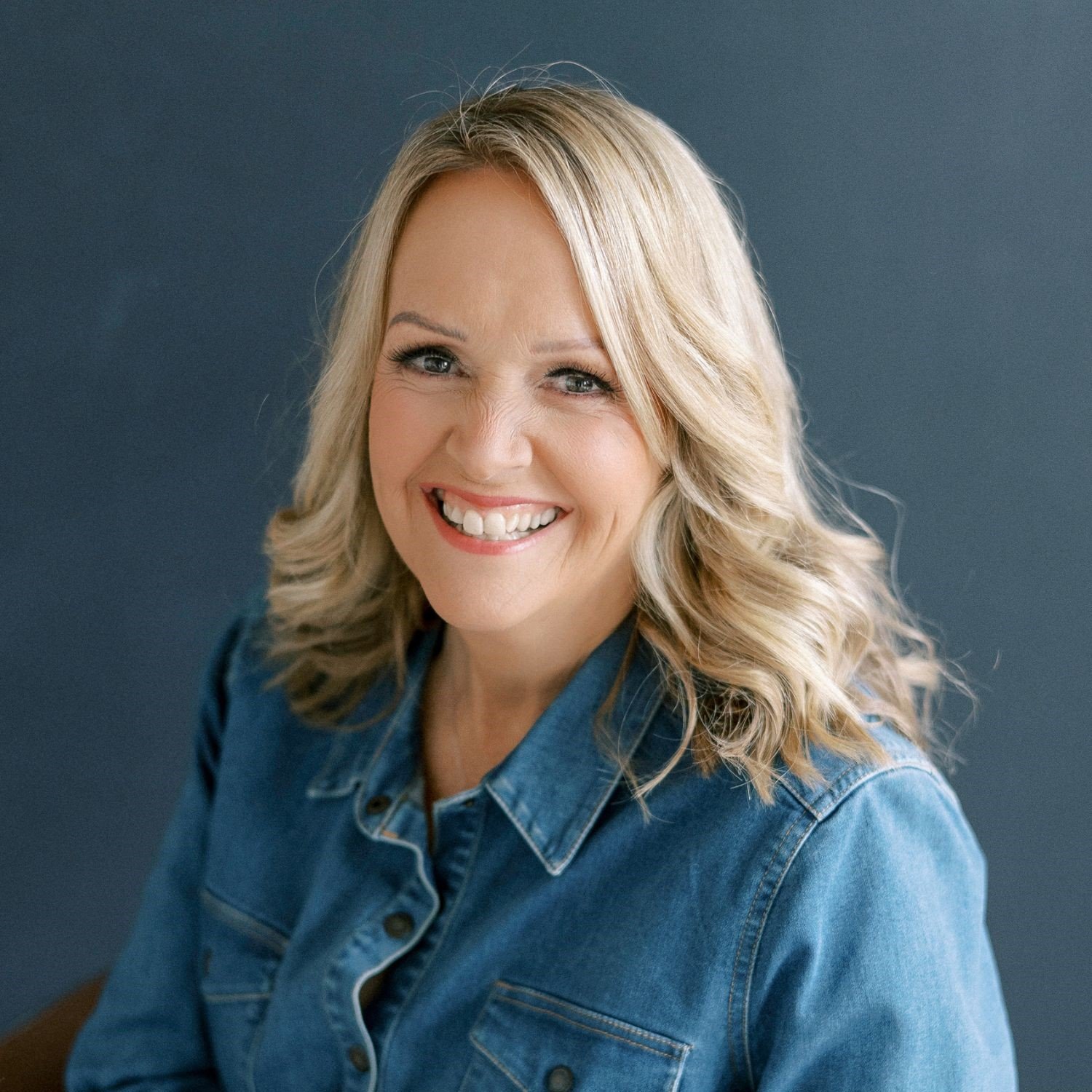 A smiling woman with blonde hair styled in loose waves, wearing a denim shirt and posing against a dark blue background.