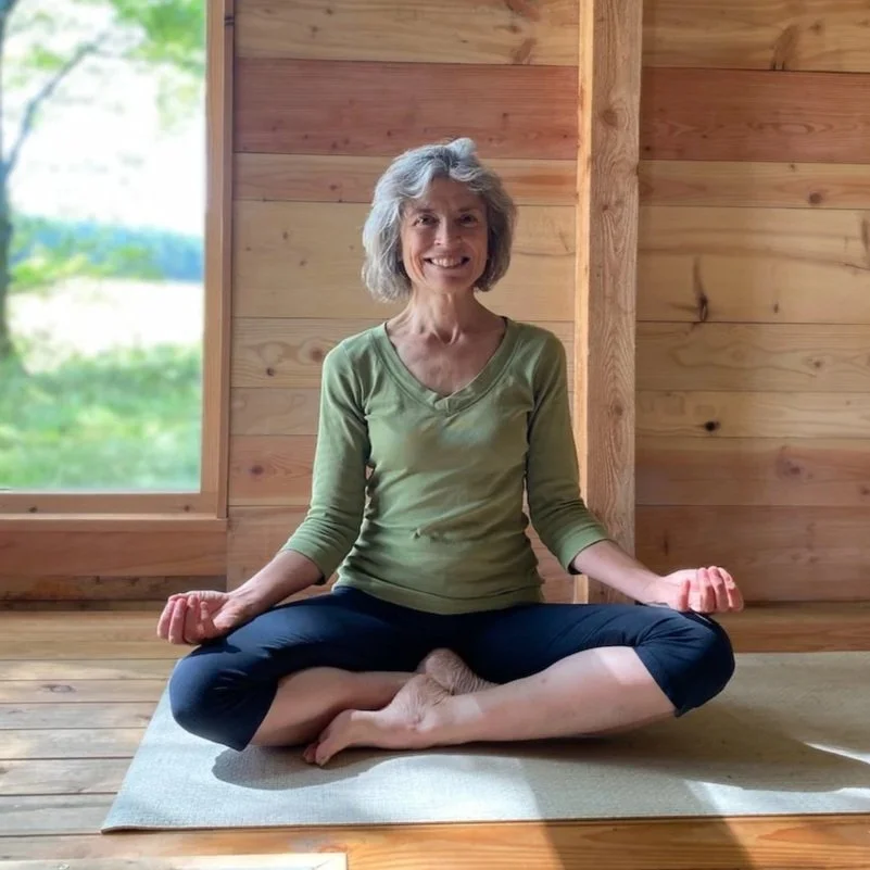 A smiling woman practicing yoga in a wooden room, seated in a cross-legged position on a mat near a window with greenery outside.