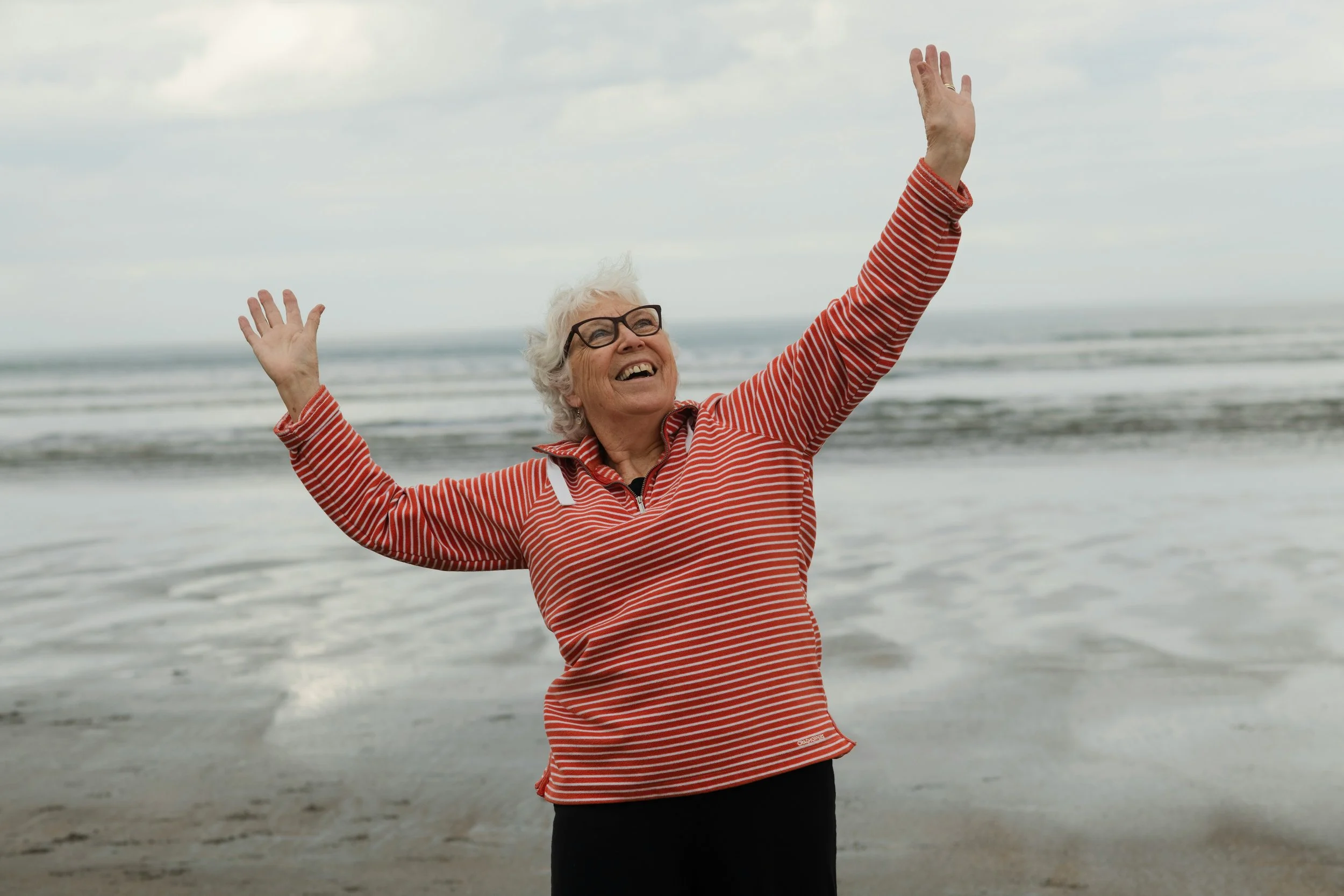 An elderly woman with glasses and curly gray hair joyfully raising her arms on a beach with ocean waves in the background.