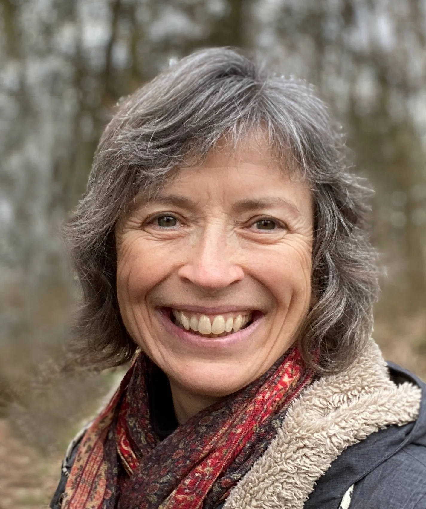 Close-up of a smiling woman with short gray hair, wearing a scarf and jacket, outdoors with blurred trees in the background.