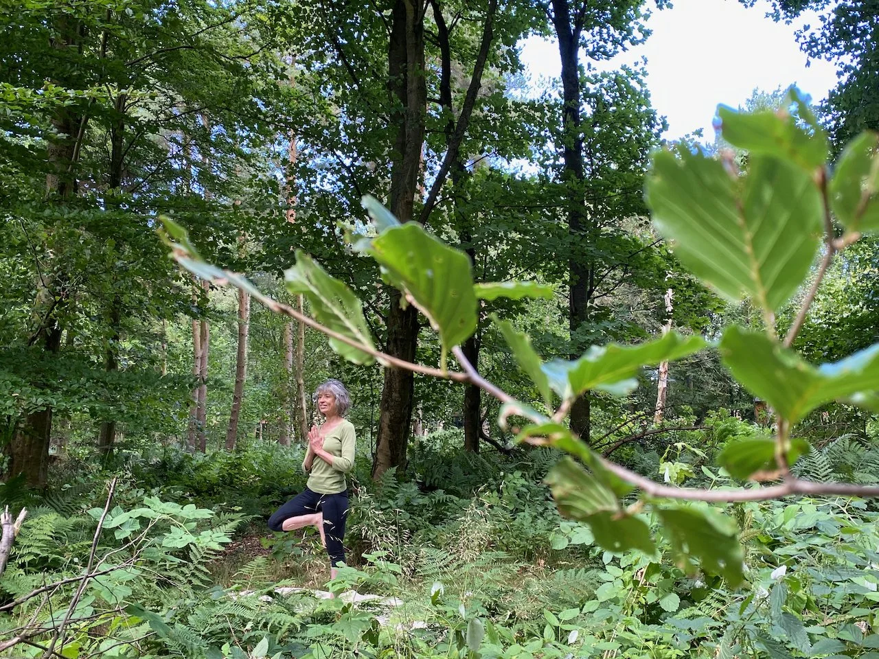 A woman practicing yoga in a forest, standing on one leg with hands in prayer position near her chest, surrounded by dense green trees and foliage.