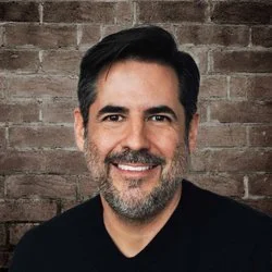 A man smiling in front of a brick wall.