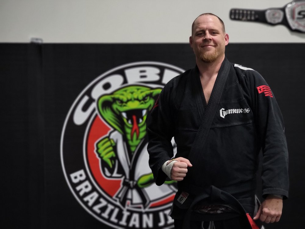 Jeff Scott, black belt, standing in front of the Cobra BJJ wall logo