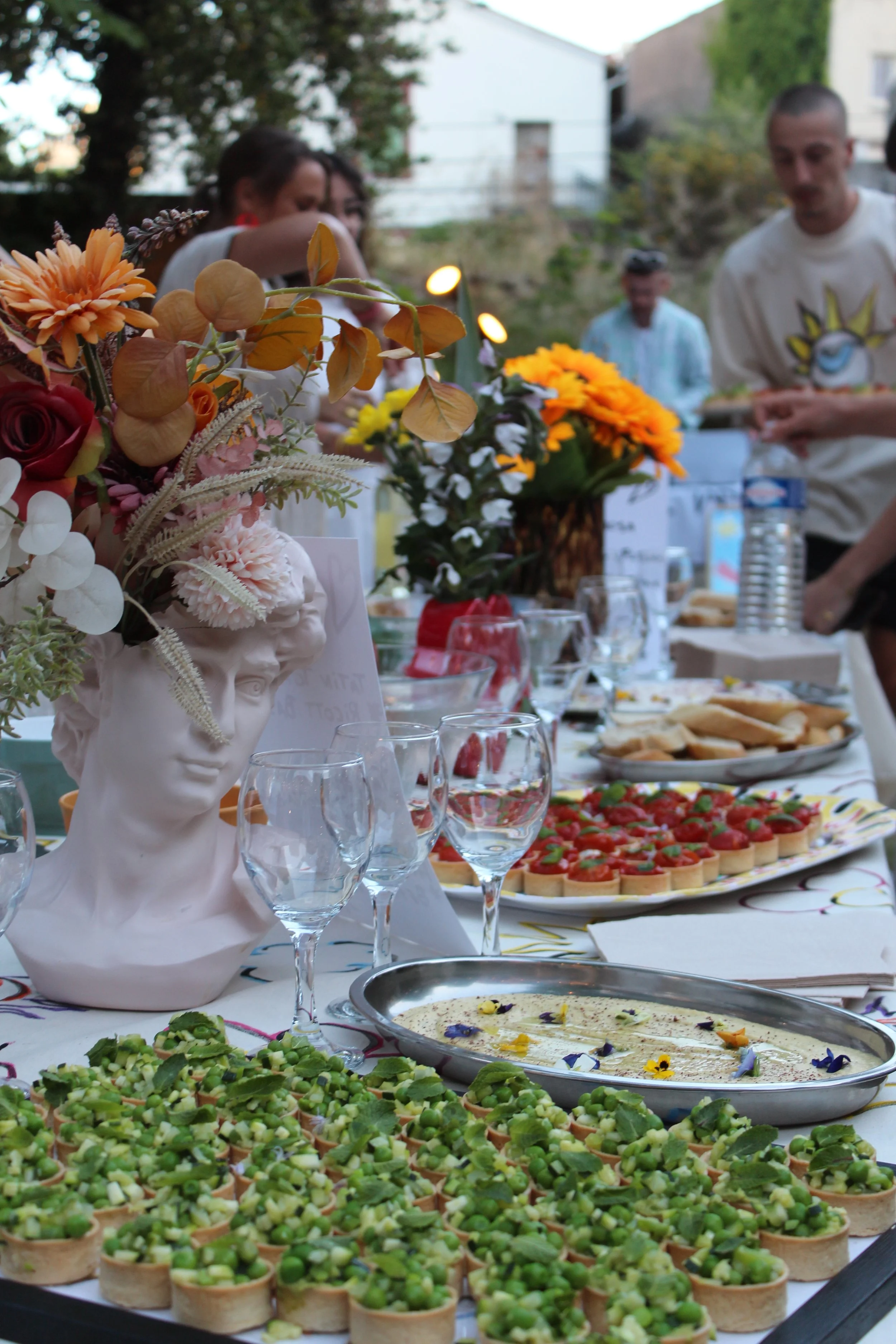 Une table de fête avec des bouquets de fleurs dans des têtes de statue, des plats apéritifs dont des tartines au caviar d'aubergine, une pizza, un plat de crème décorée de fleurs comestibles, et des verres à vin, lors d'une réunion en plein air.