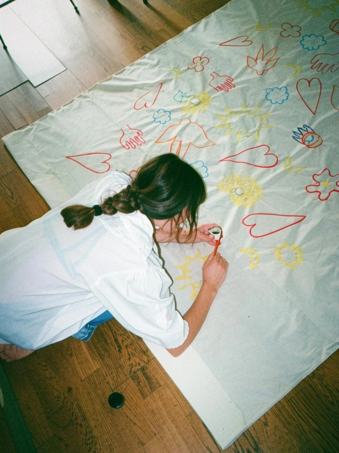Une femme dessine sur une grande feuille blanche posée sur une table en bois, avec des dessins colorés de cœurs, soleils, nuages, et autres formes.