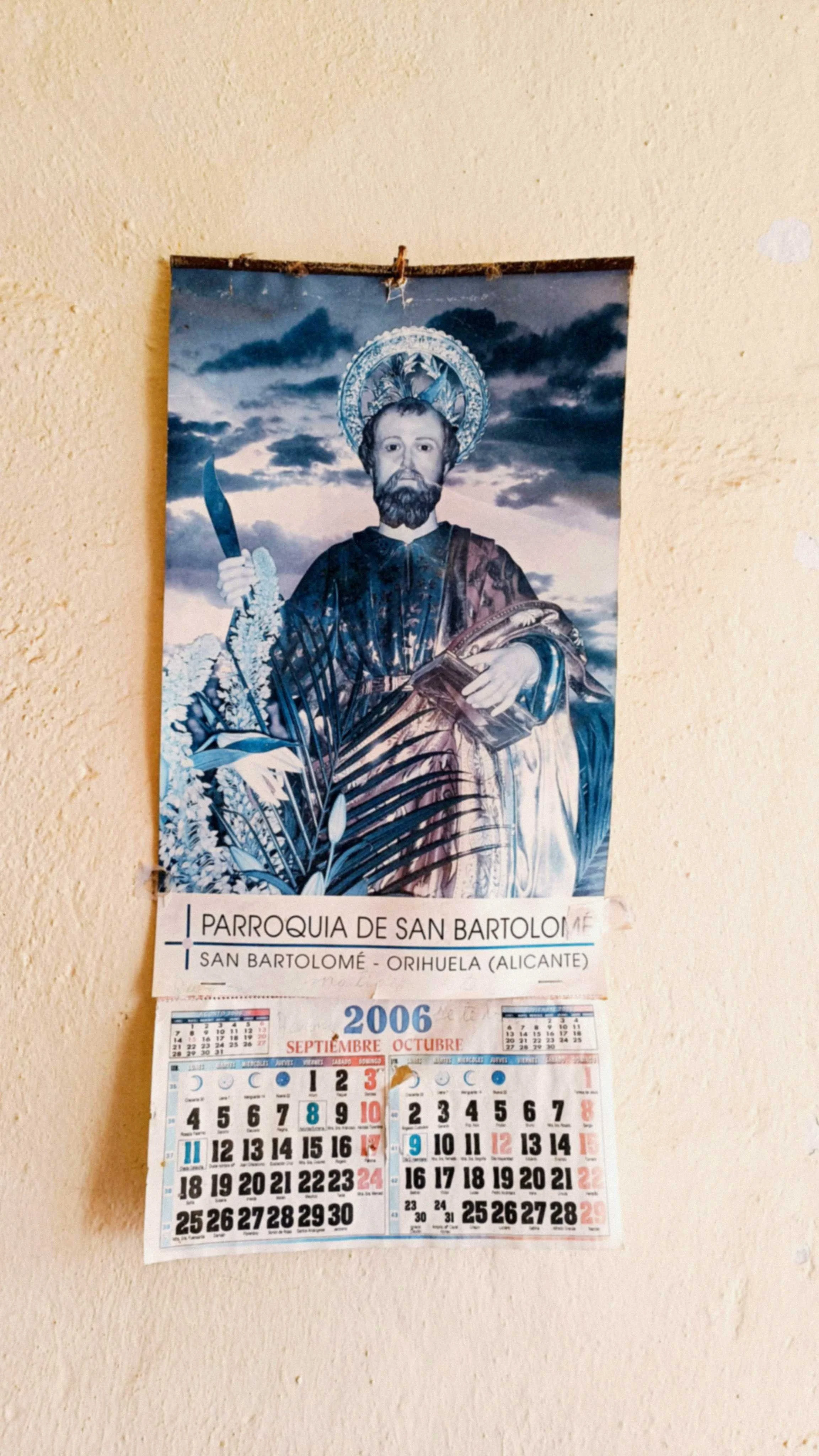 Affiche religieuse représentant saint avec halo, barbe, tenue sombre, tenant un poignard, avec un ciel orageux en arrière-plan, sur un mur beige, avec calendrier mensuel de septembre-octobre 2006