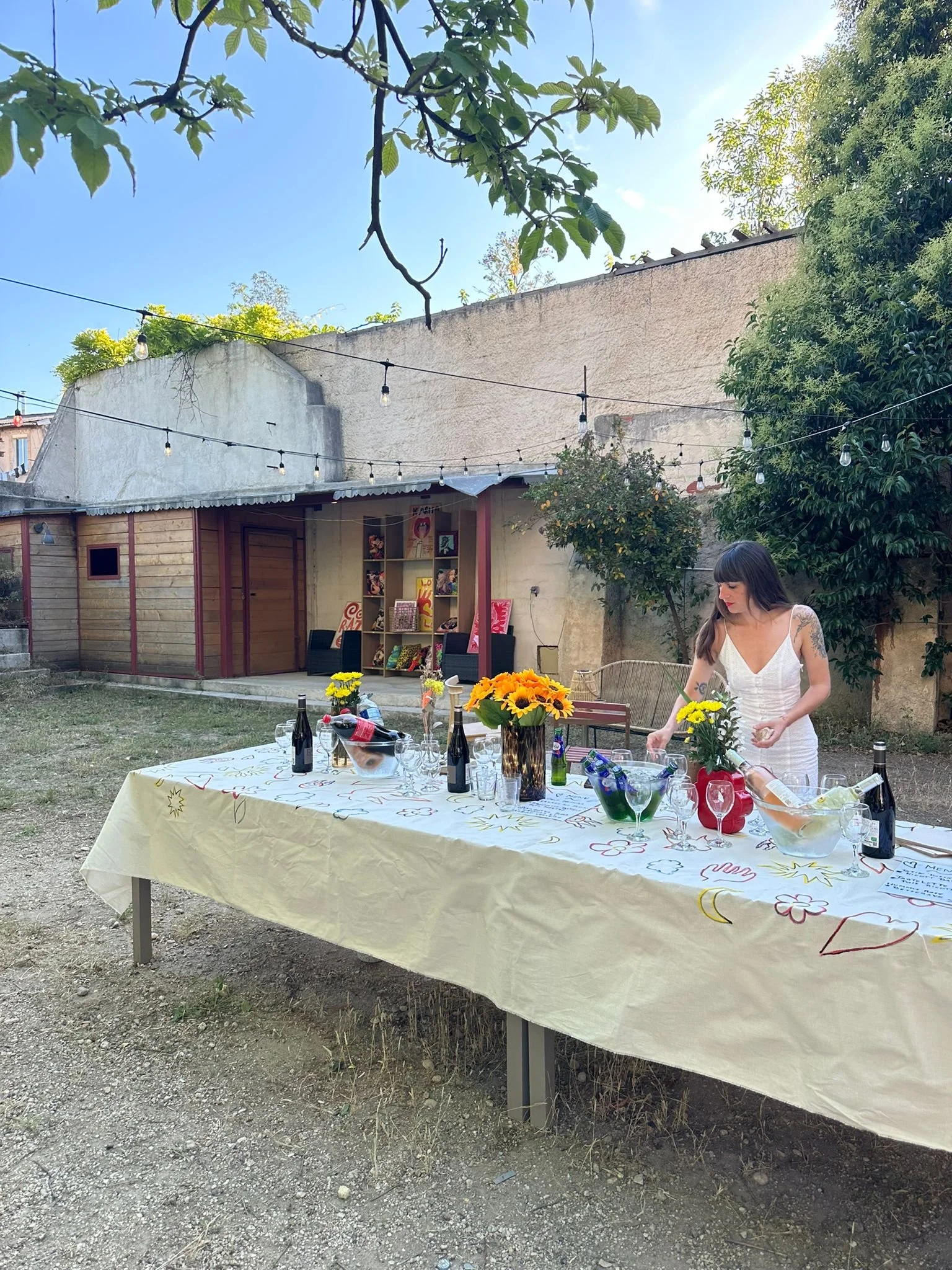 Une scène en extérieur avec une table décorée avec des fleurs, des bouteilles de vin, et des verres. Une jeune femme portant une robe blanche se tient à côté de la table. Il y a des lampes suspendues au-dessus, un mur en arrière-plan avec des étagères et des œuvres d'art affichées. L’environnement est verdoyant avec des arbres lus très grands et un ciel clair.
