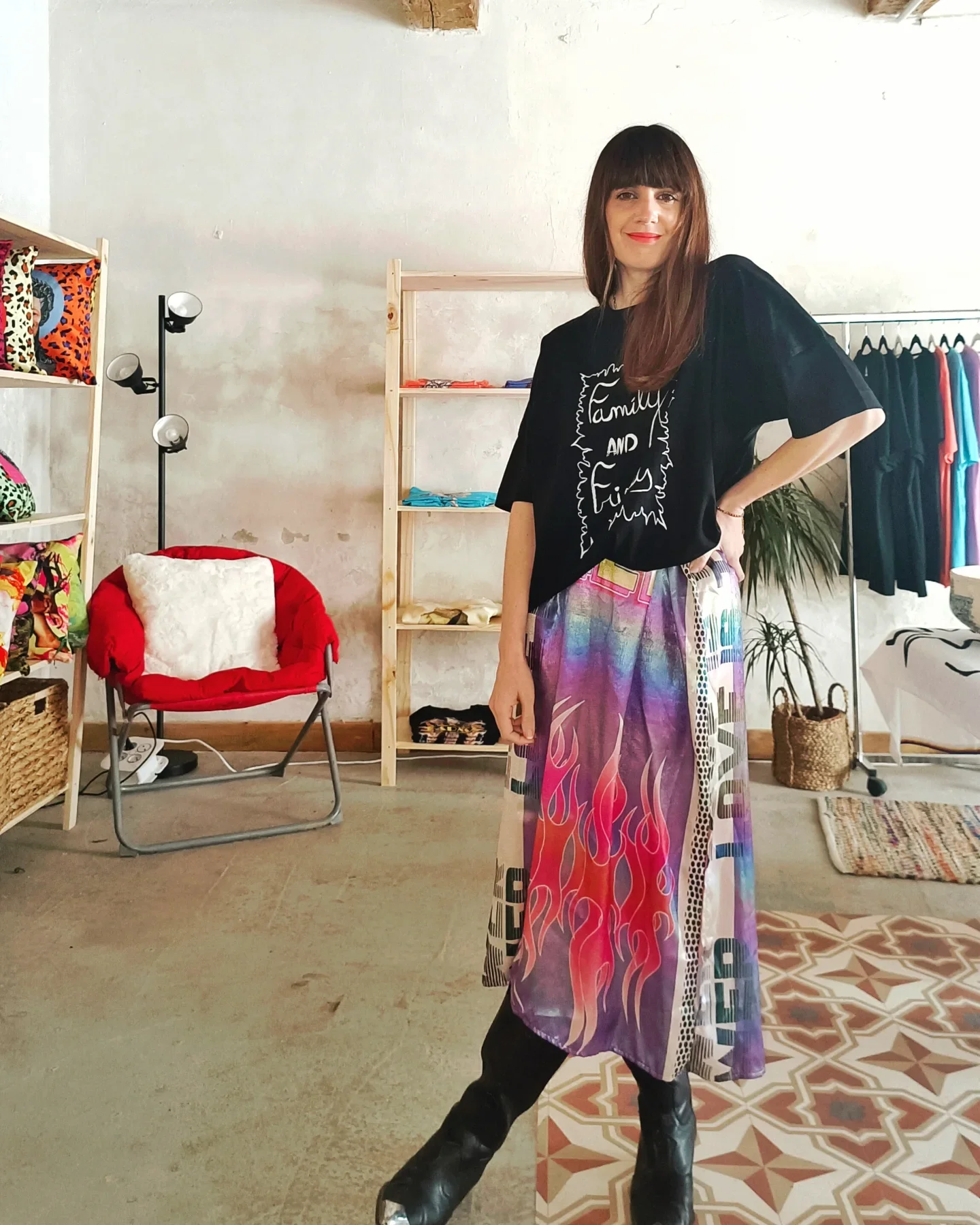 Une femme debout dans un espace intérieur, portant un t-shirt noir avec un dessin et un texte, et une jupe colorée avec un motif de flammes. Elle a les cheveux bruns et longs, avec des franges, et sourit à la caméra. En arrière-plan, il y a des étagères en bois, un fauteuil rouge avec un coussin blanc, un lampadaire moderne, ainsi que des vêtements suspendus et des plantes.