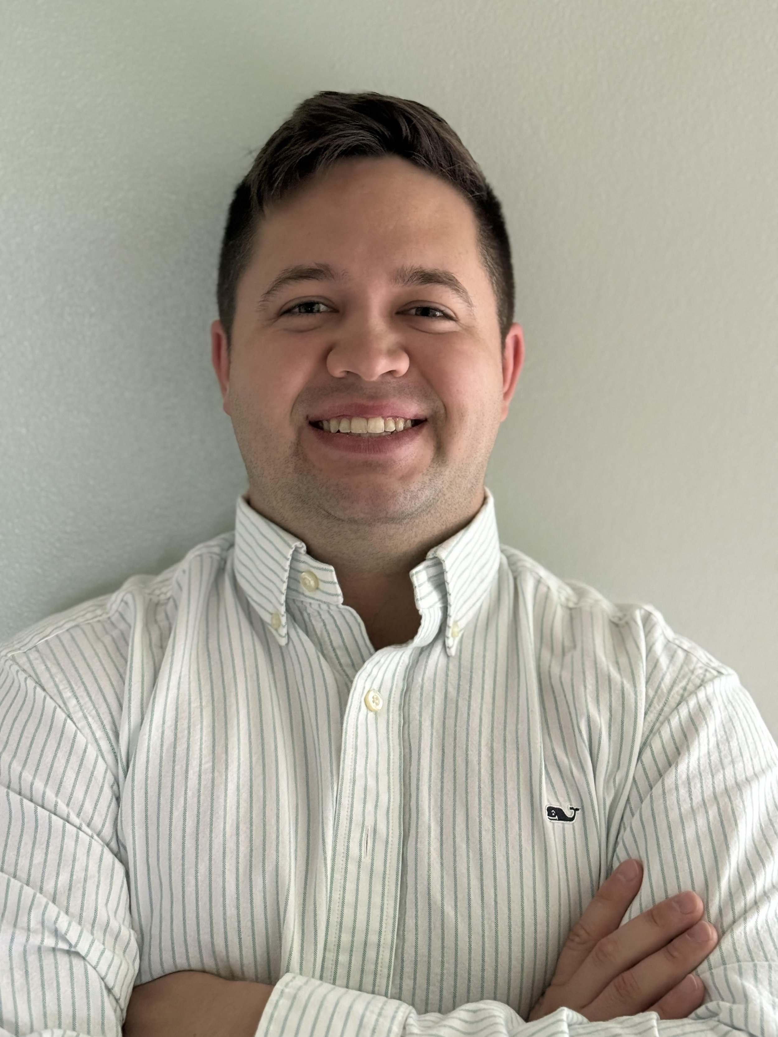 A young man with short dark hair smiling, standing against a light-colored wall, wearing a white striped button-up shirt with the collar buttoned, arms crossed.