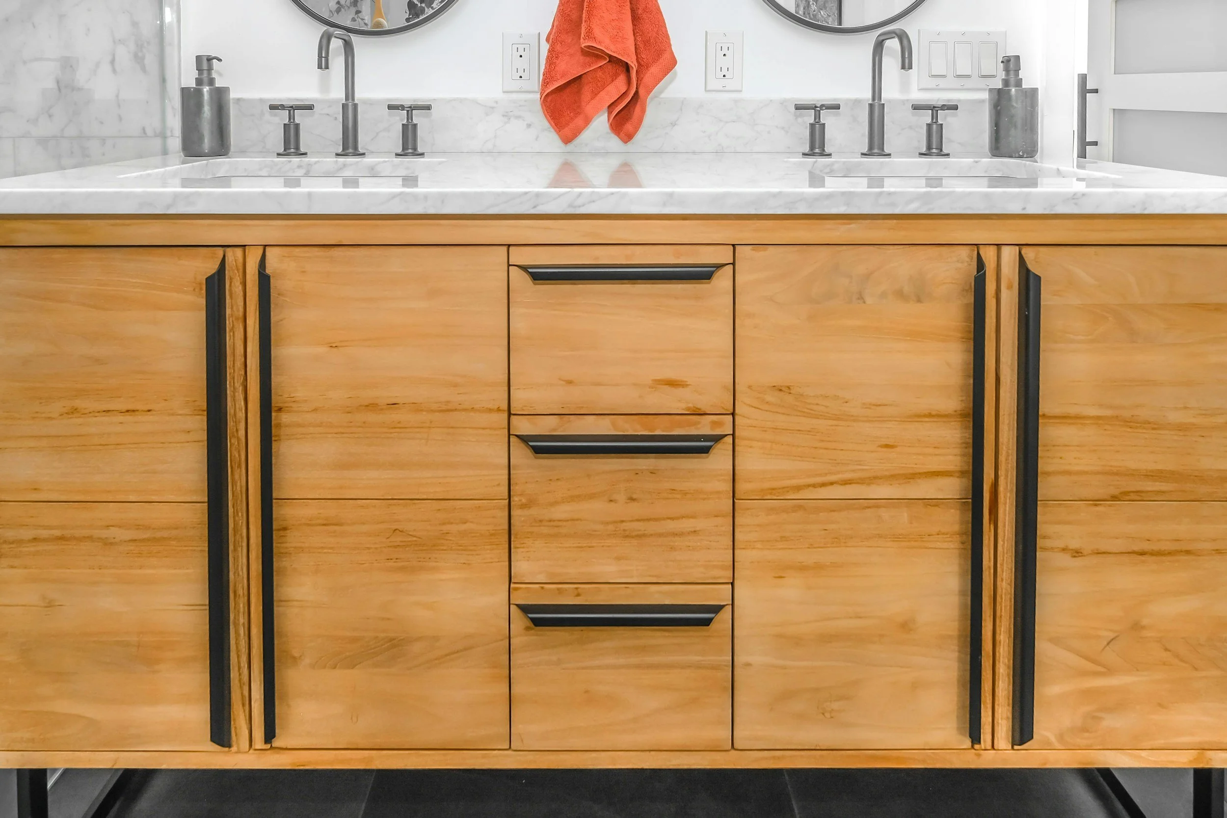Wooden bathroom vanity with marble countertop, two sinks, and black handles, orange towel hanging on a wall, and electrical outlets