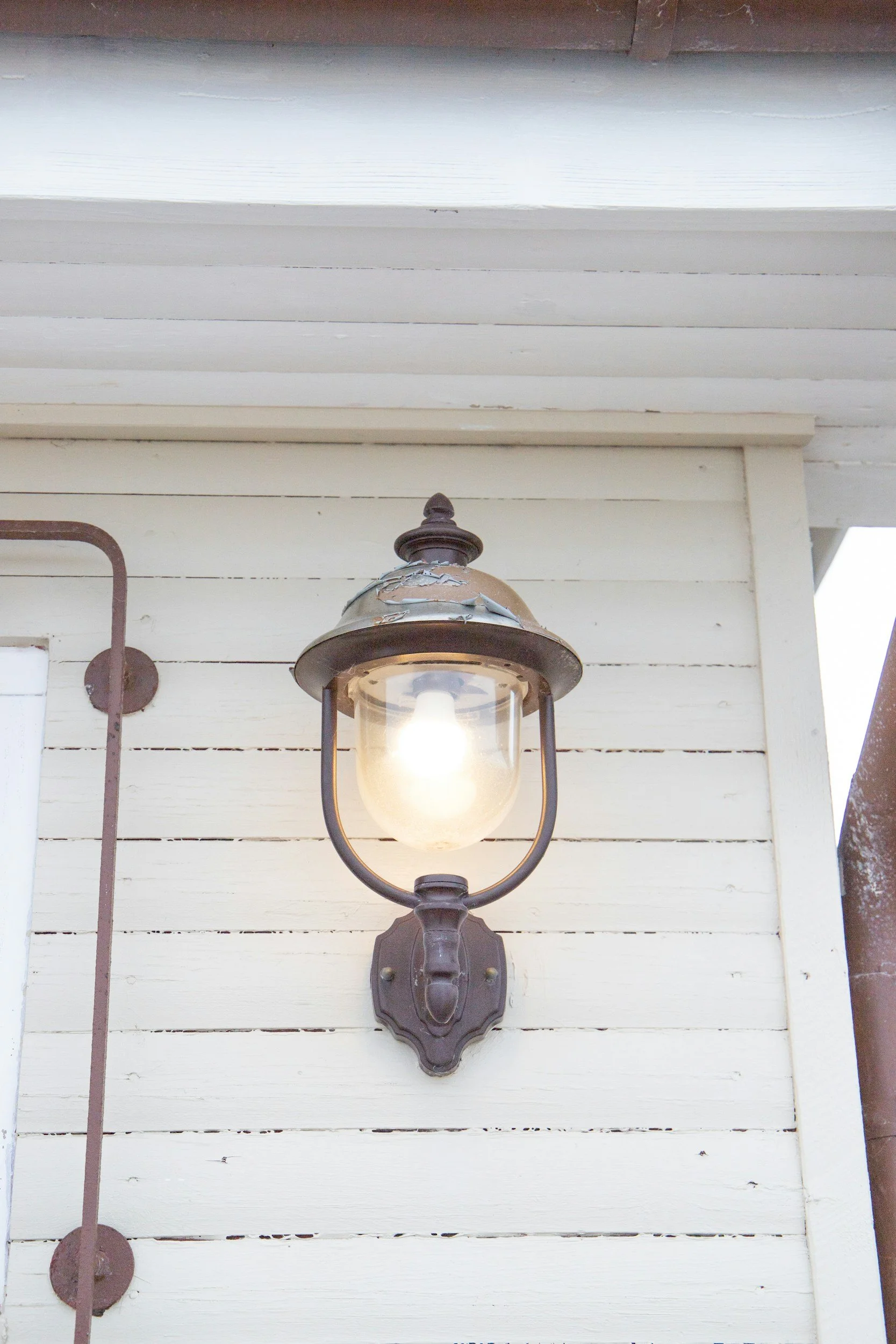 Outdoor vintage style wall-mounted lantern with a lit bulb, attached to white wooden siding of a building.