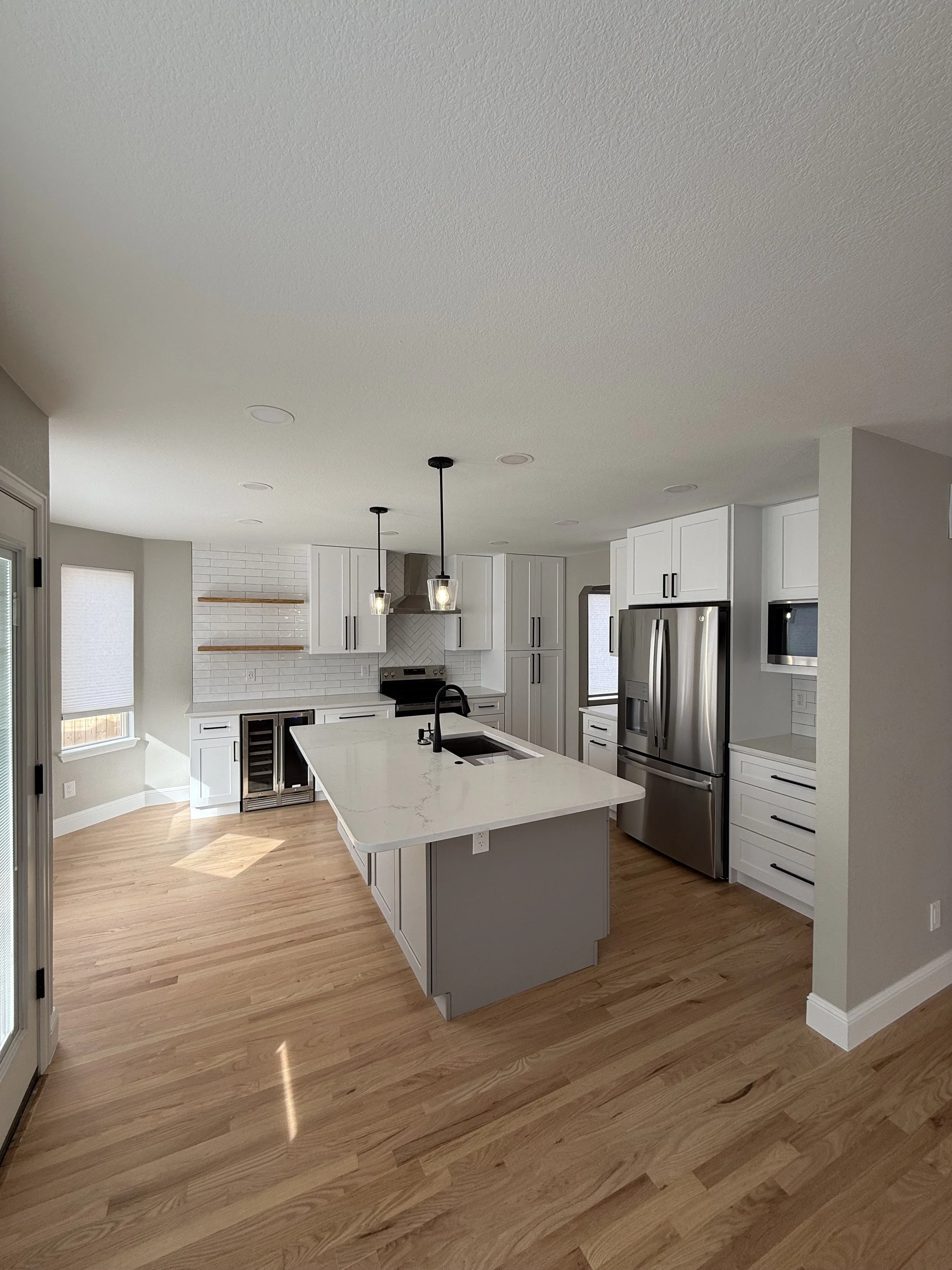 A modern kitchen with white cabinets, stainless steel appliances, a white marble island, pendant lights, and light wood flooring.