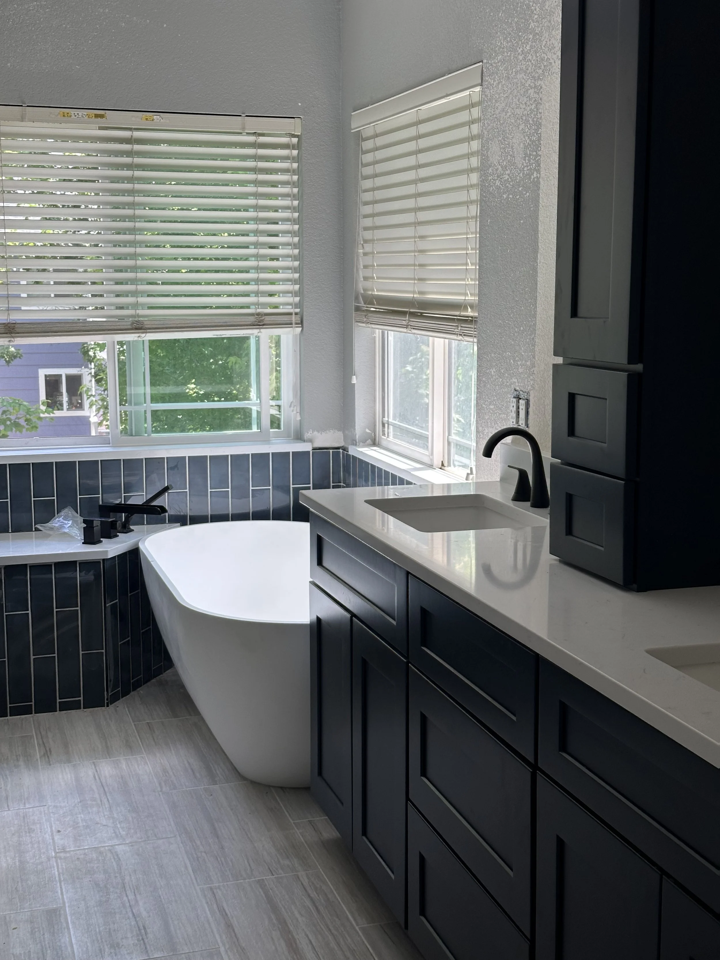 Modern bathroom with a freestanding bathtub near windows with white blinds, dark blue cabinets, white countertop with a sink, and light wood floor tiles.