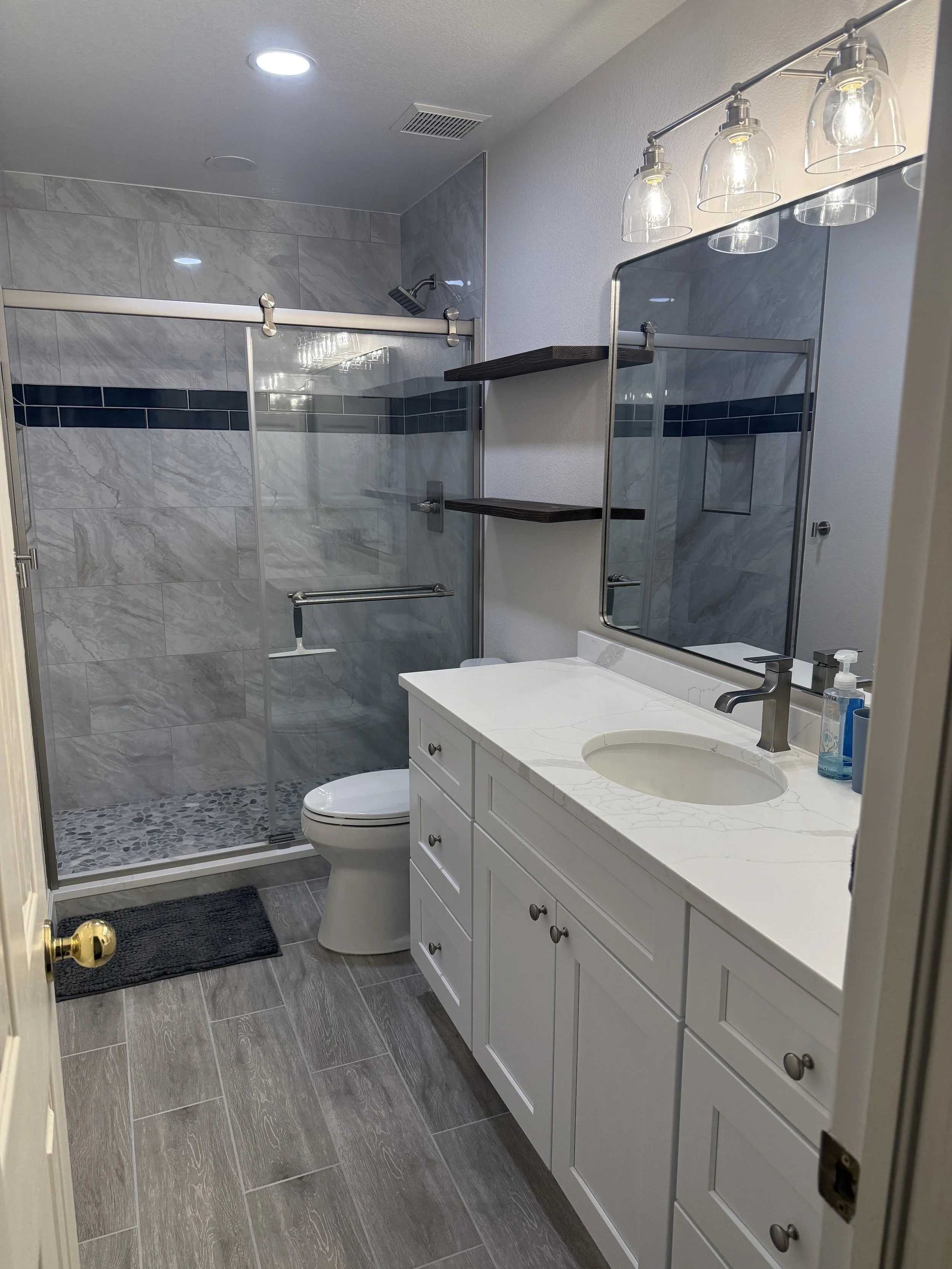 A modern bathroom with a glass-enclosed shower, a white vanity with a marble countertop, a large mirror, a wooden shelf, a soap dispenser, and wall-mounted lighting fixtures.