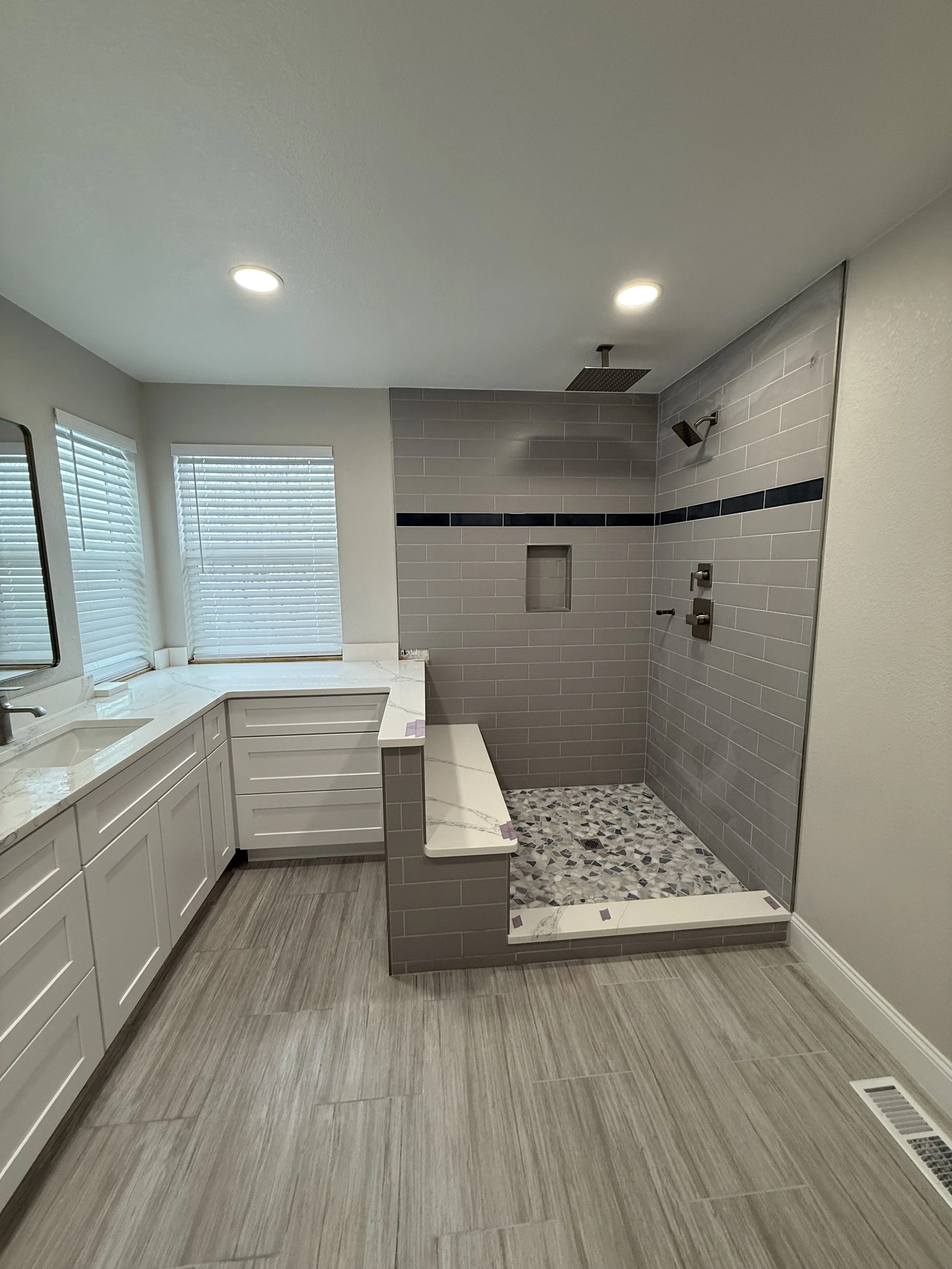Modern bathroom with a walk-in shower featuring gray subway tiles, a pebble floor, and a built-in niche. There are two windows with white blinds, white vanity with a marble countertop, and beige wood-look tile flooring.