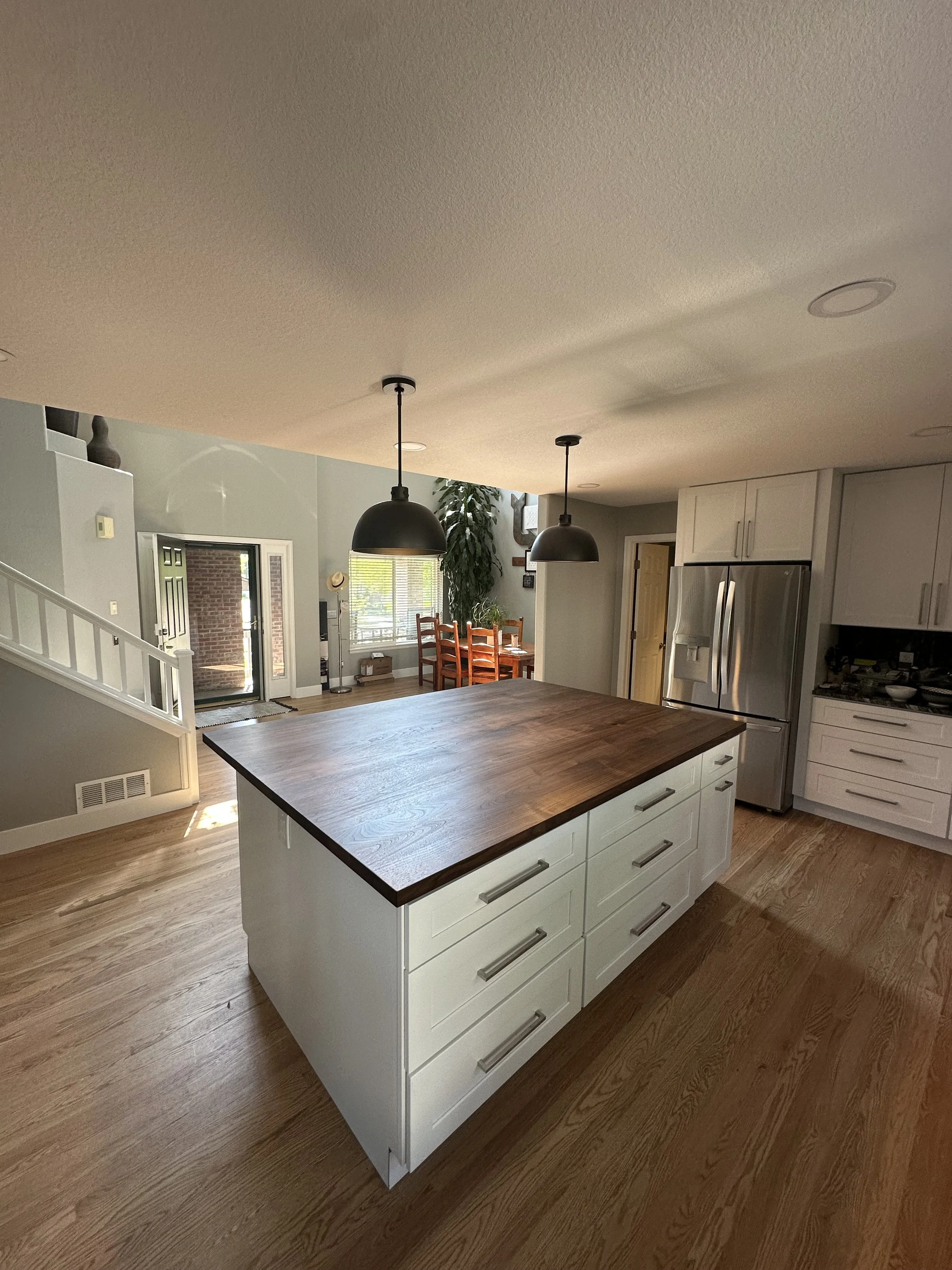 Modern kitchen with white cabinets, a large wooden island, stainless steel refrigerator, and a dining area with wooden tables and chairs, featuring hardwood floors and pendant lighting.