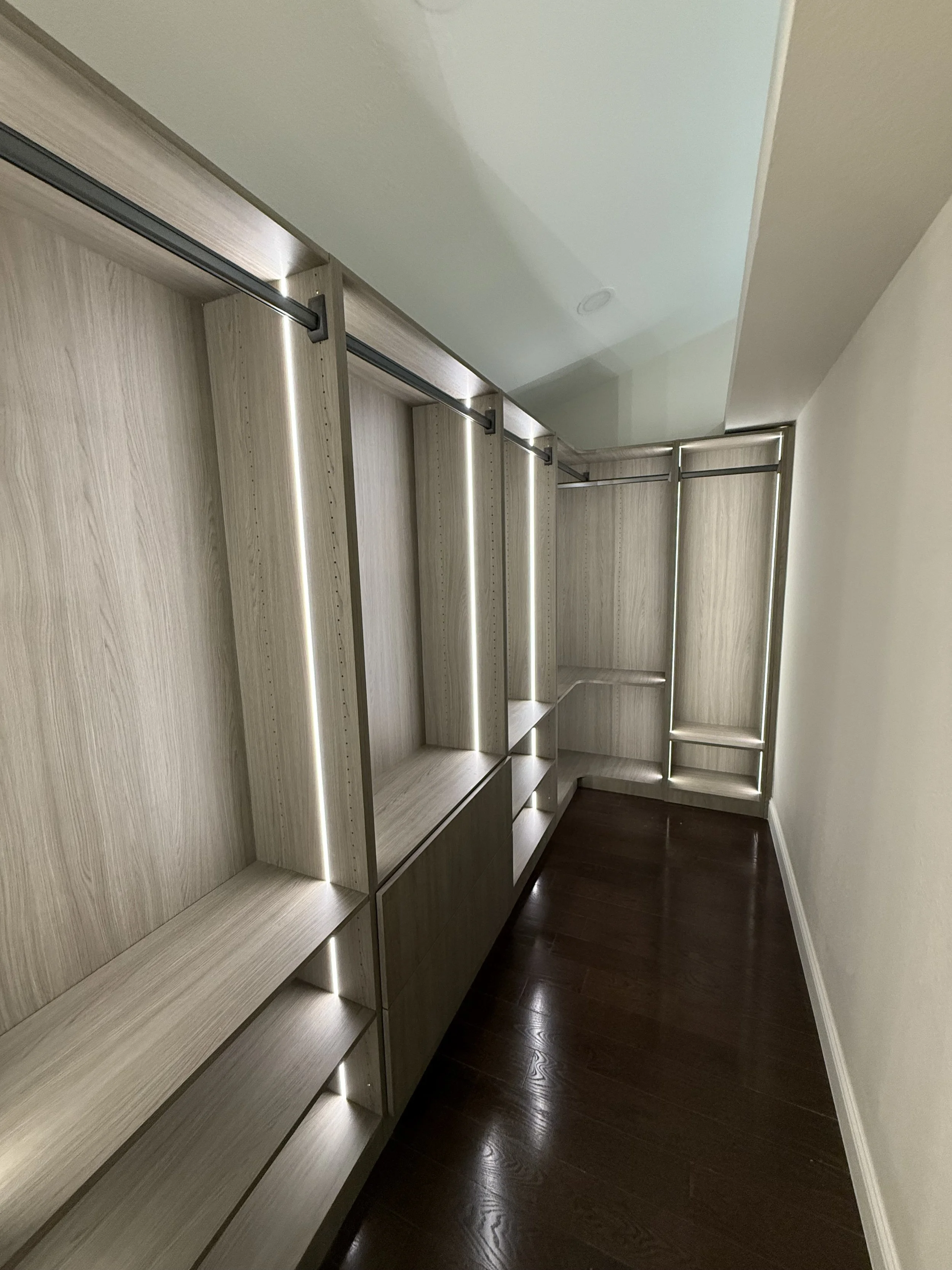 Interior view of a modern walk-in closet featuring light wood shelving units with LED strip lighting and dark hardwood floors.