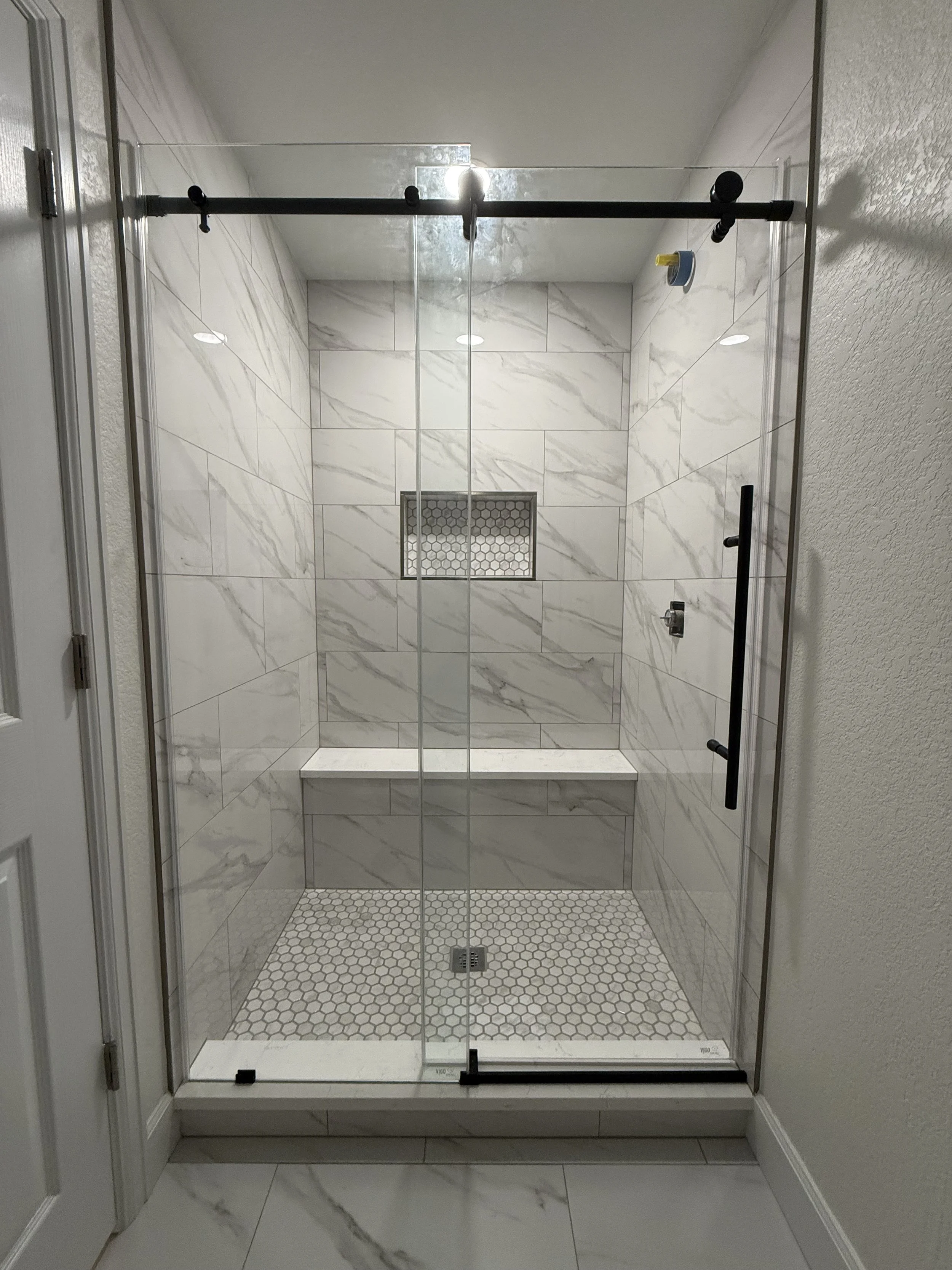 Modern walk-in shower with glass door, white marble tiles, a built-in bench, a small hexagonal tile floor, and a drain in the center.