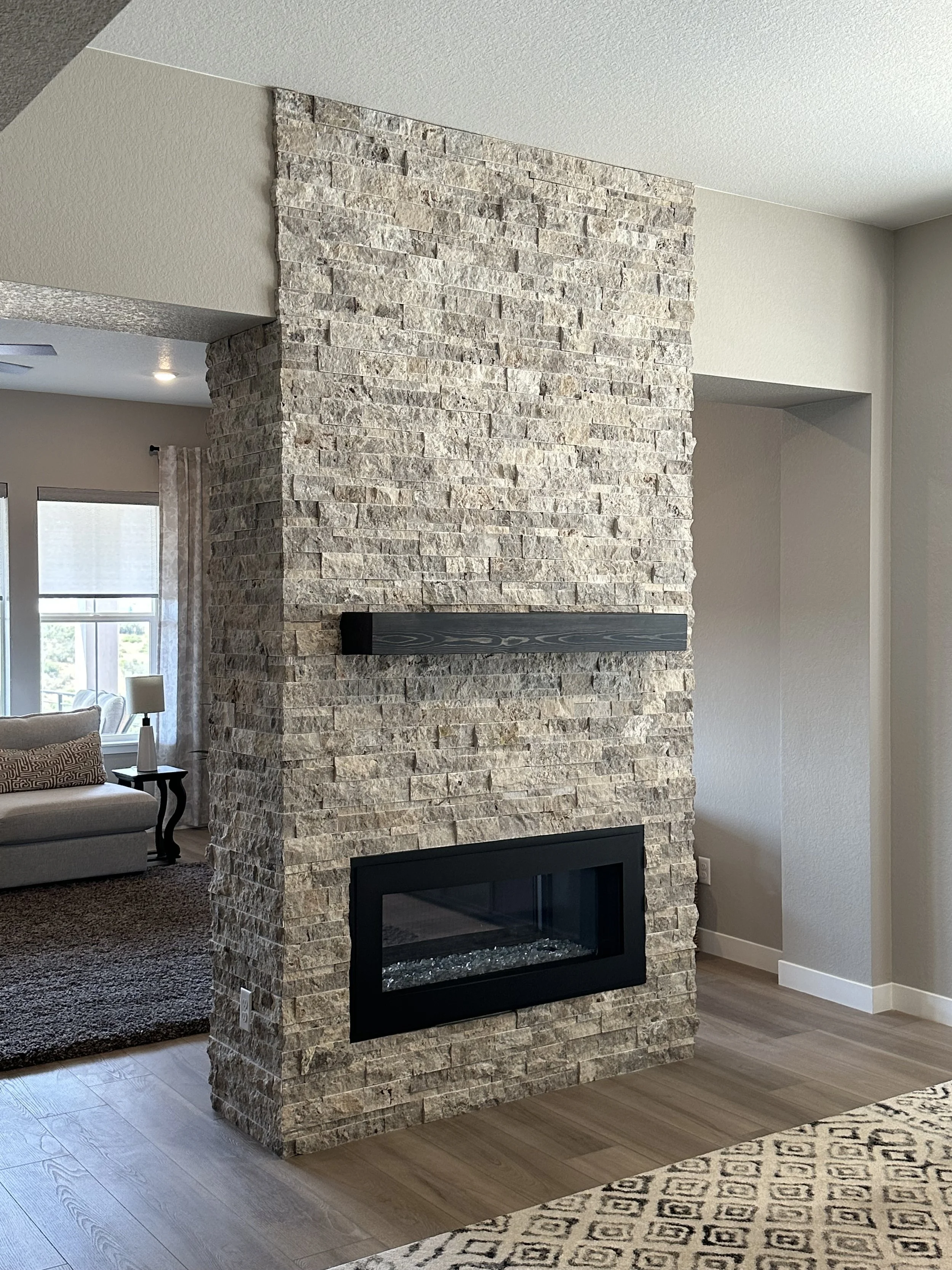 Photo of a stone fireplace with a black metal trim in a living room, next to a beige wall and a window with curtains.