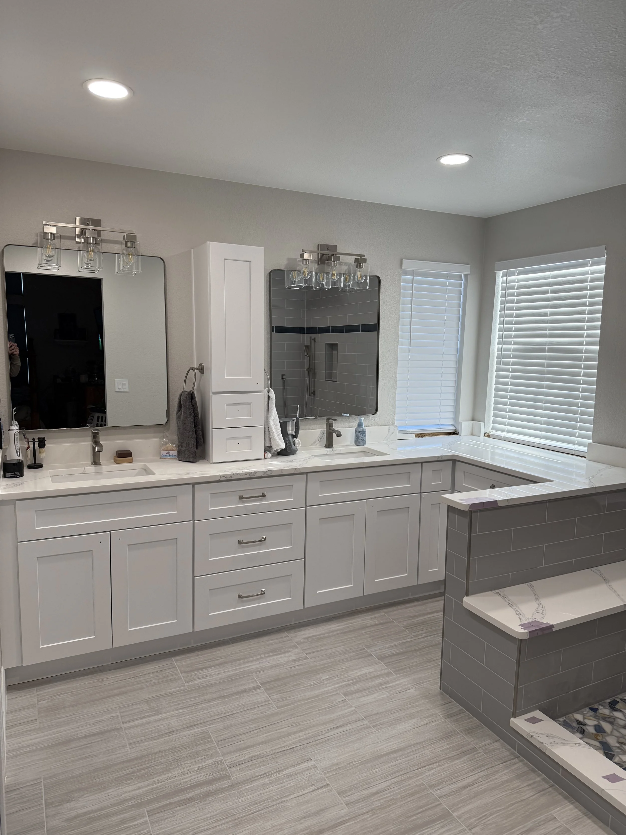A modern bathroom vanity area with white cabinets, two large mirrors, and light fixtures on the ceiling. There are two sinks, one with a soap dispenser and another with a towel hanging. The wall behind the vanity has a shower area visible through one of the mirrors. There are three windows with white blinds and a small section of gray tiled wall near the bottom right corner.