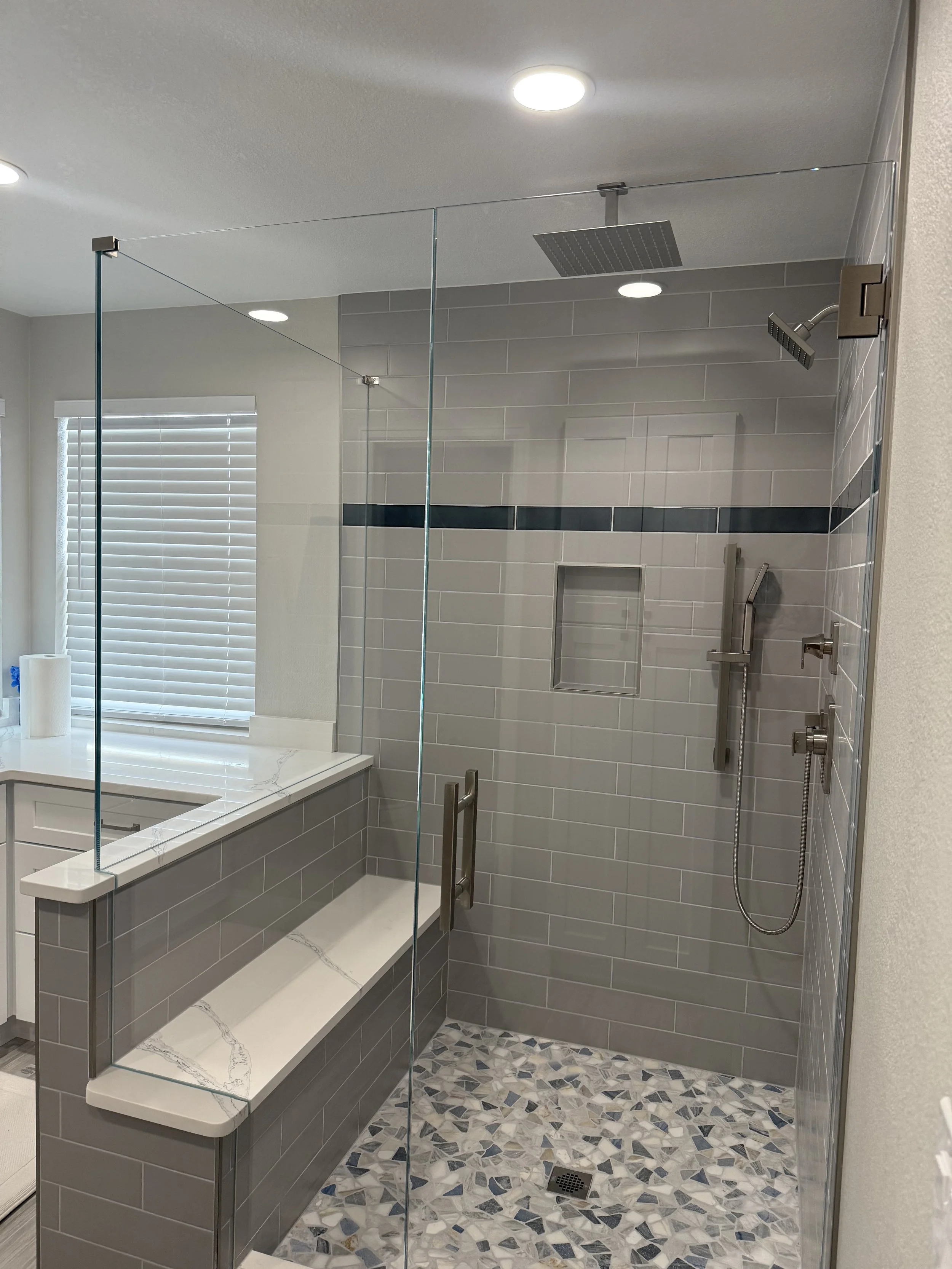 Modern bathroom with a walk-in shower featuring gray tile walls, rainfall showerheads, casual seating with a white marble top, and a mosaic tile floor.