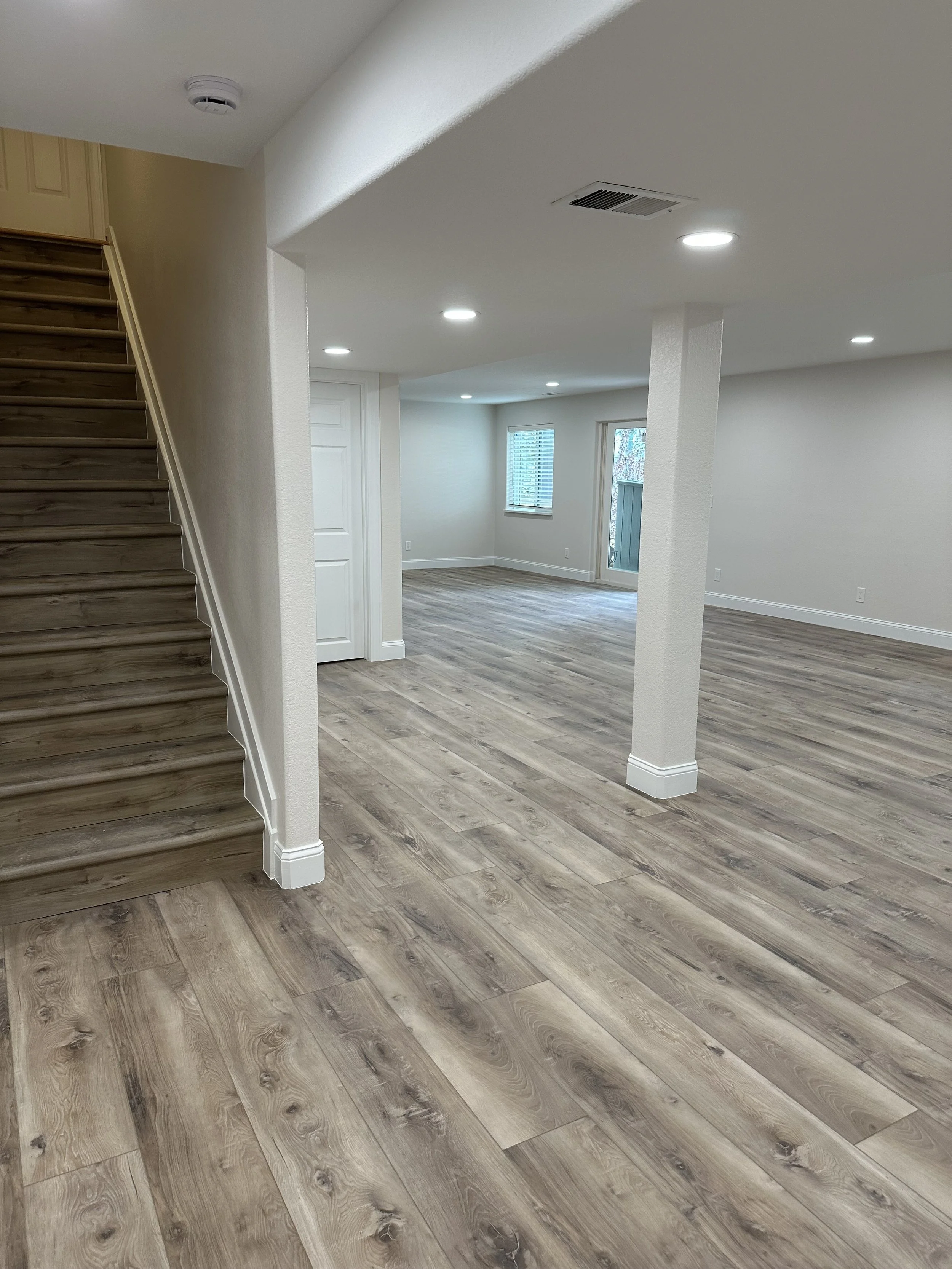 Empty living room with light-colored wood flooring, white walls, multiple windows, and a staircase with wooden steps on the left.