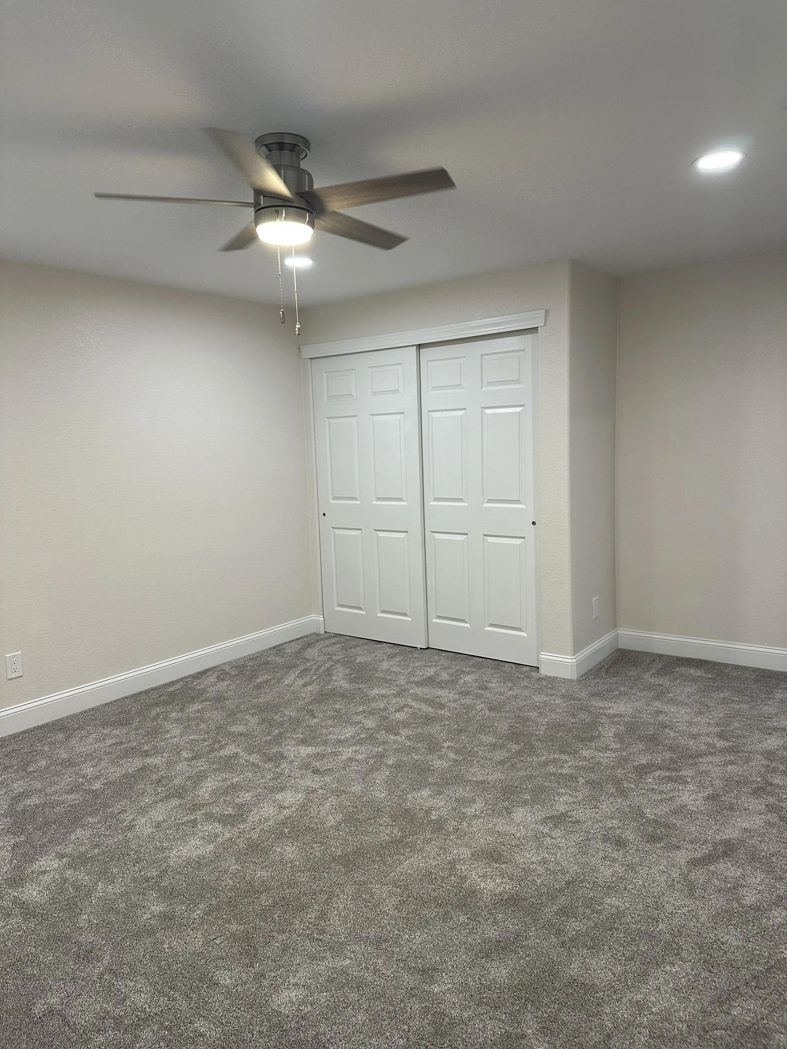 Empty room with beige walls, gray carpet, white closet doors, ceiling fan with light, and ceiling recessed lights.