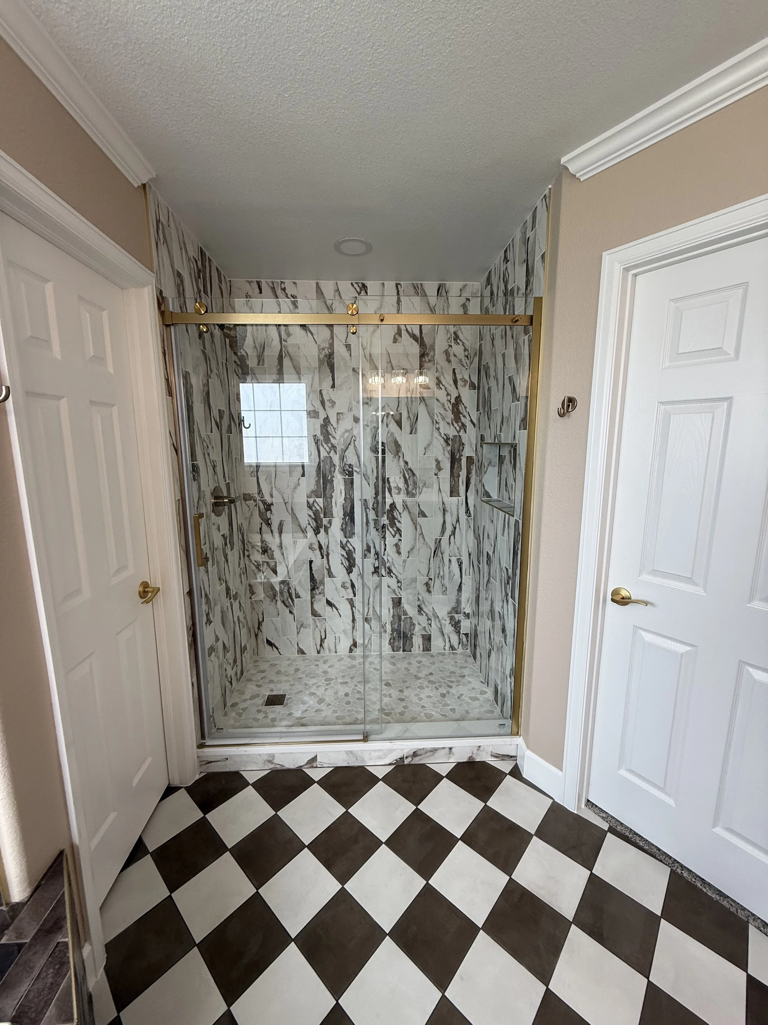 View of a bathroom shower with sliding glass doors, marble-tiled walls, and a window. The floor has a black and white checkered tile pattern.