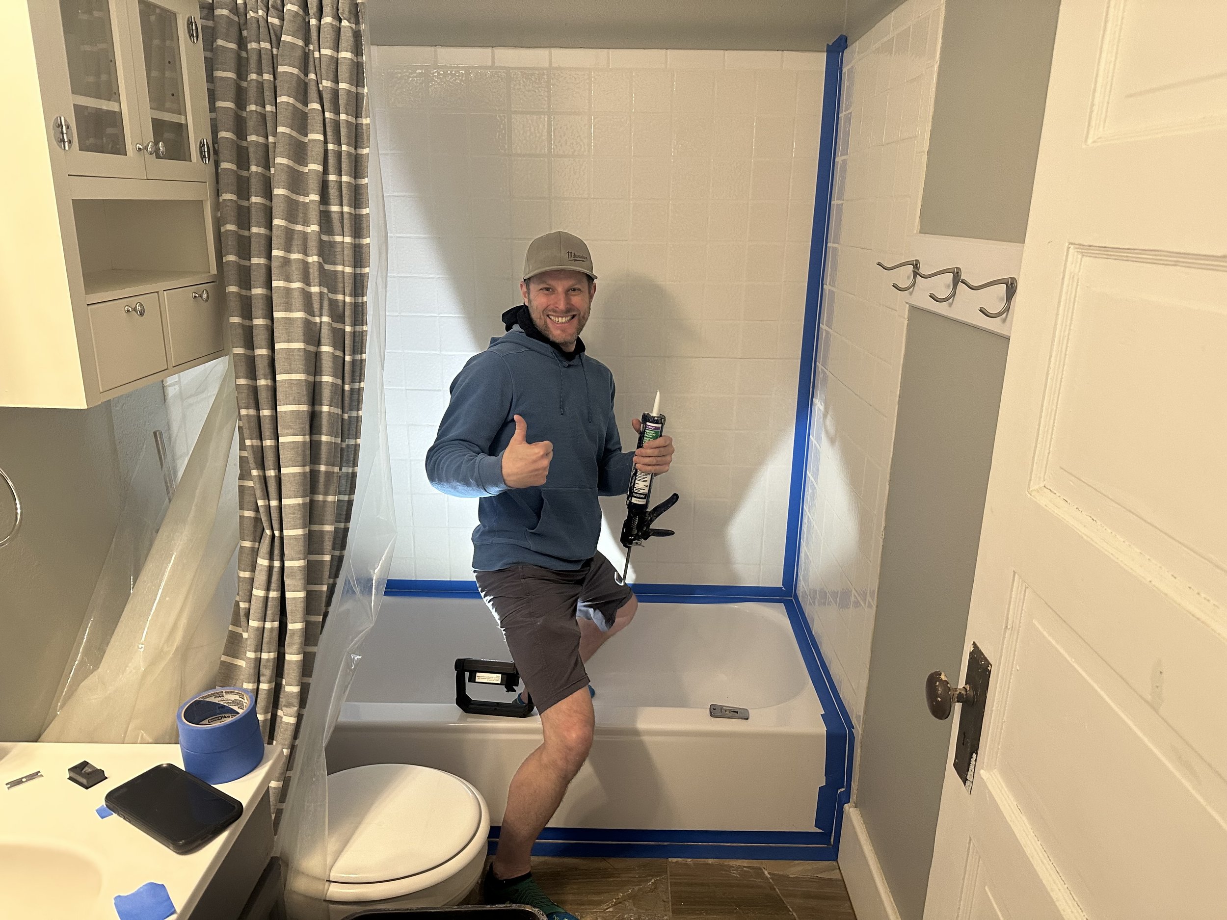 A man standing in a bathroom, giving a thumbs-up, holding a caulking tube, near a shower with blue painter's tape on the edges. The bathroom has a cabinet, a toilet with a drumstick on top, and a partially opened door with hooks.