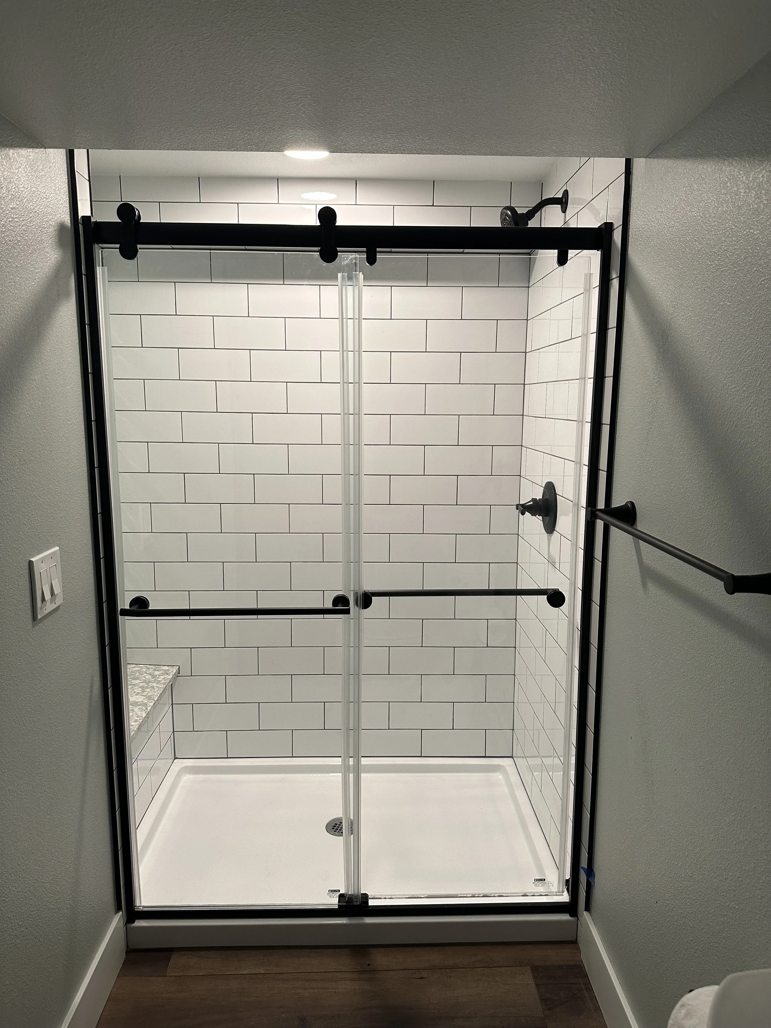 A walk-in shower with sliding glass doors, tiled walls with white subway tiles, black hardware, a built-in bench, and a rainfall showerhead.