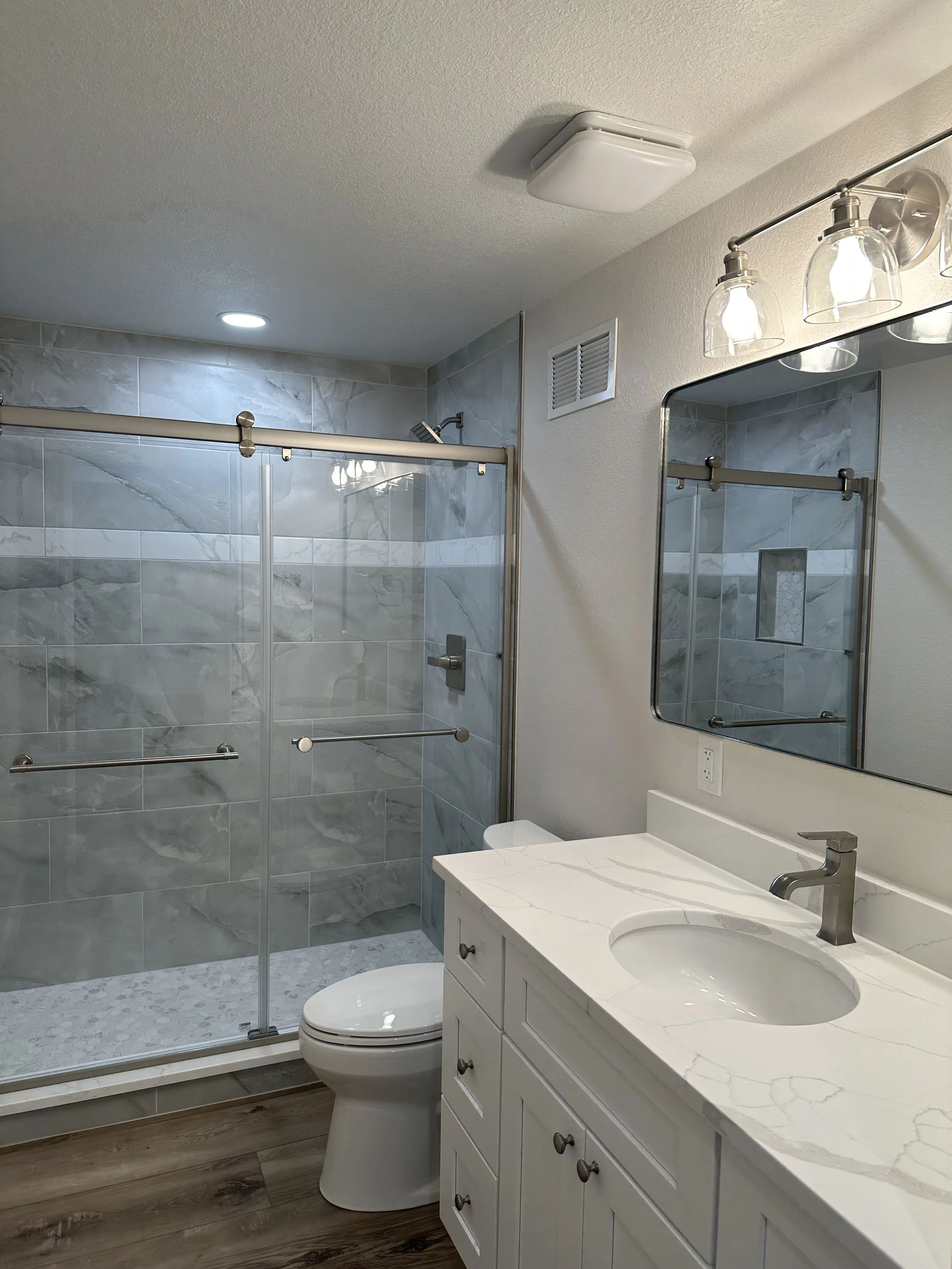 Modern bathroom with a walk-in glass shower, white vanity with a marble countertop, and a large mirror with three light fixtures above.