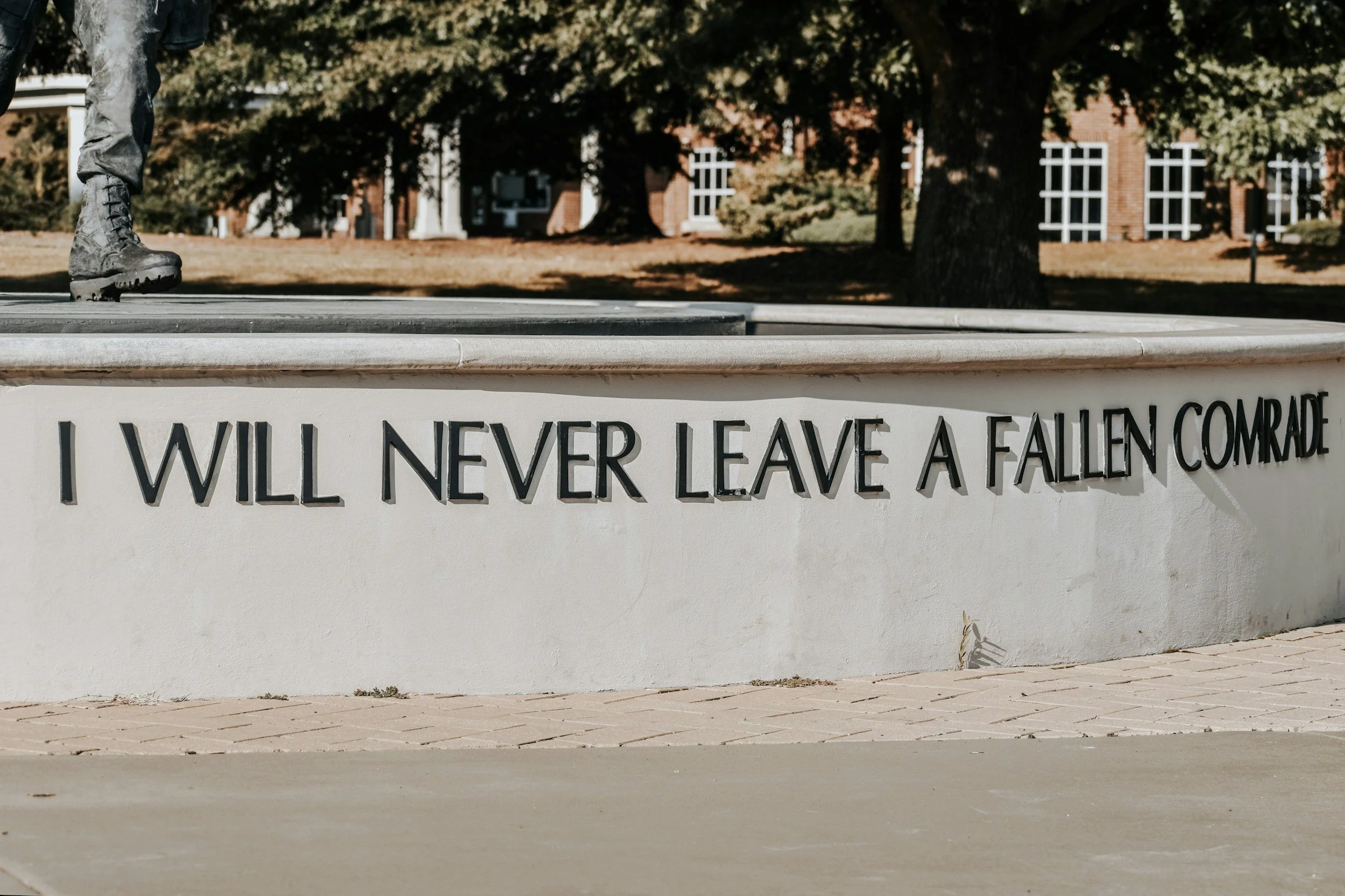 Close-up of a white stone monument with a statue of a soldier's legs in combat boots on top. The monument features engraved text that reads, 'I WILL NEVER LEAVE A FALLEN COMRADE.' In the background, there are trees and a brick building with white-framed windows.