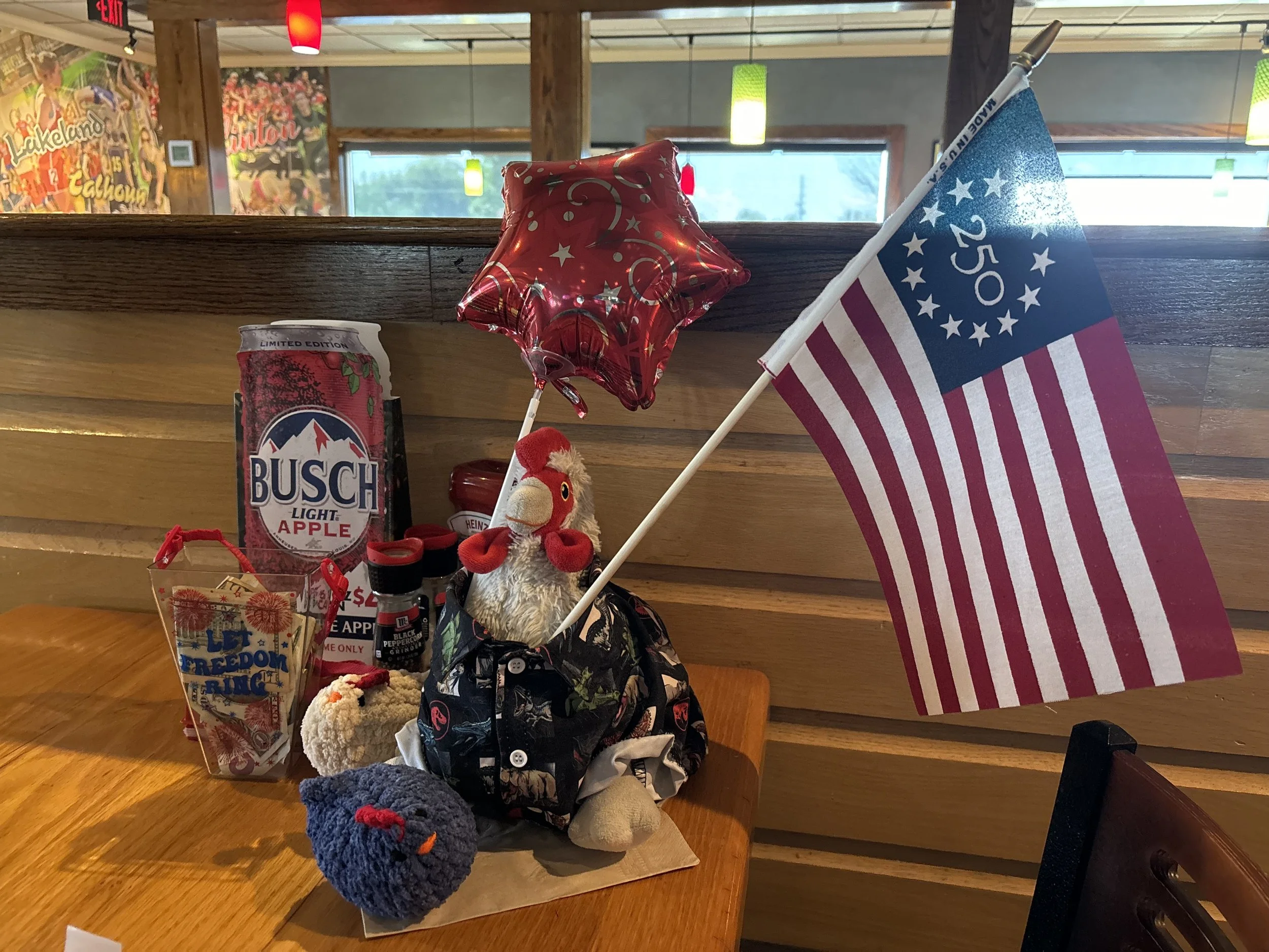 Table decorated with patriotic items including a small American flag, a star-shaped balloon, a plush turkey toy, a can of Busch Light Apple beer, and various small plush toys. Background features a mural, windows, and hanging lights in a cozy indoor setting.
