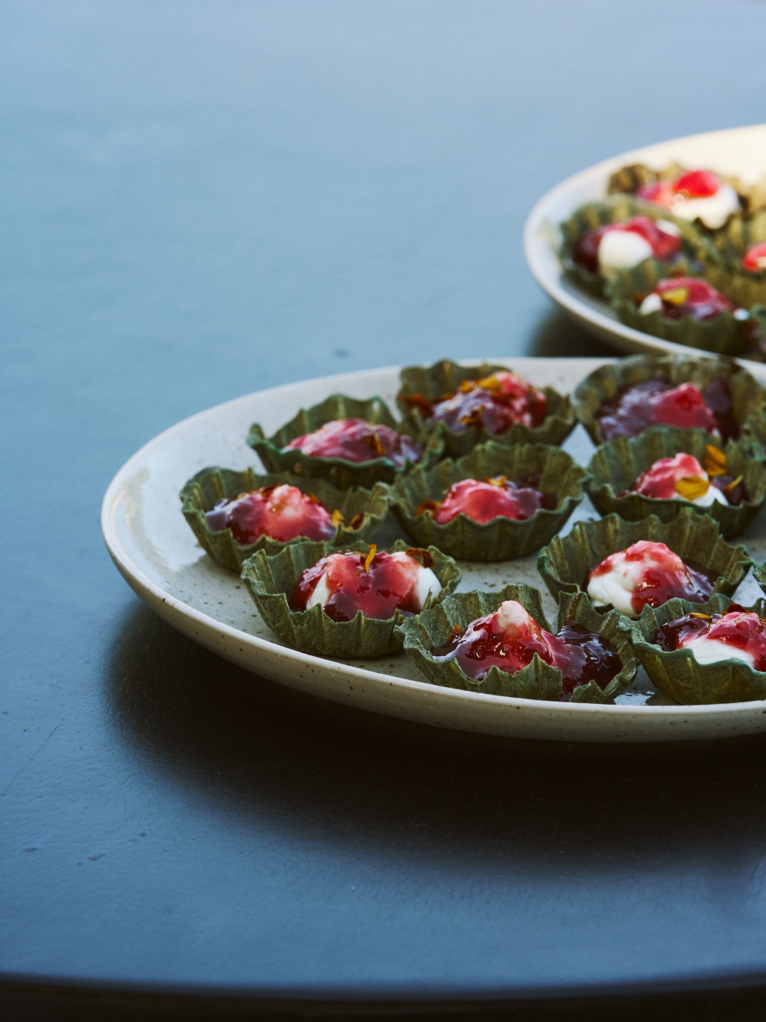 Two white plates, each with small desserts served in green edible cups, topped with a red jelly and a dollop of white cream, set on a blue surface.