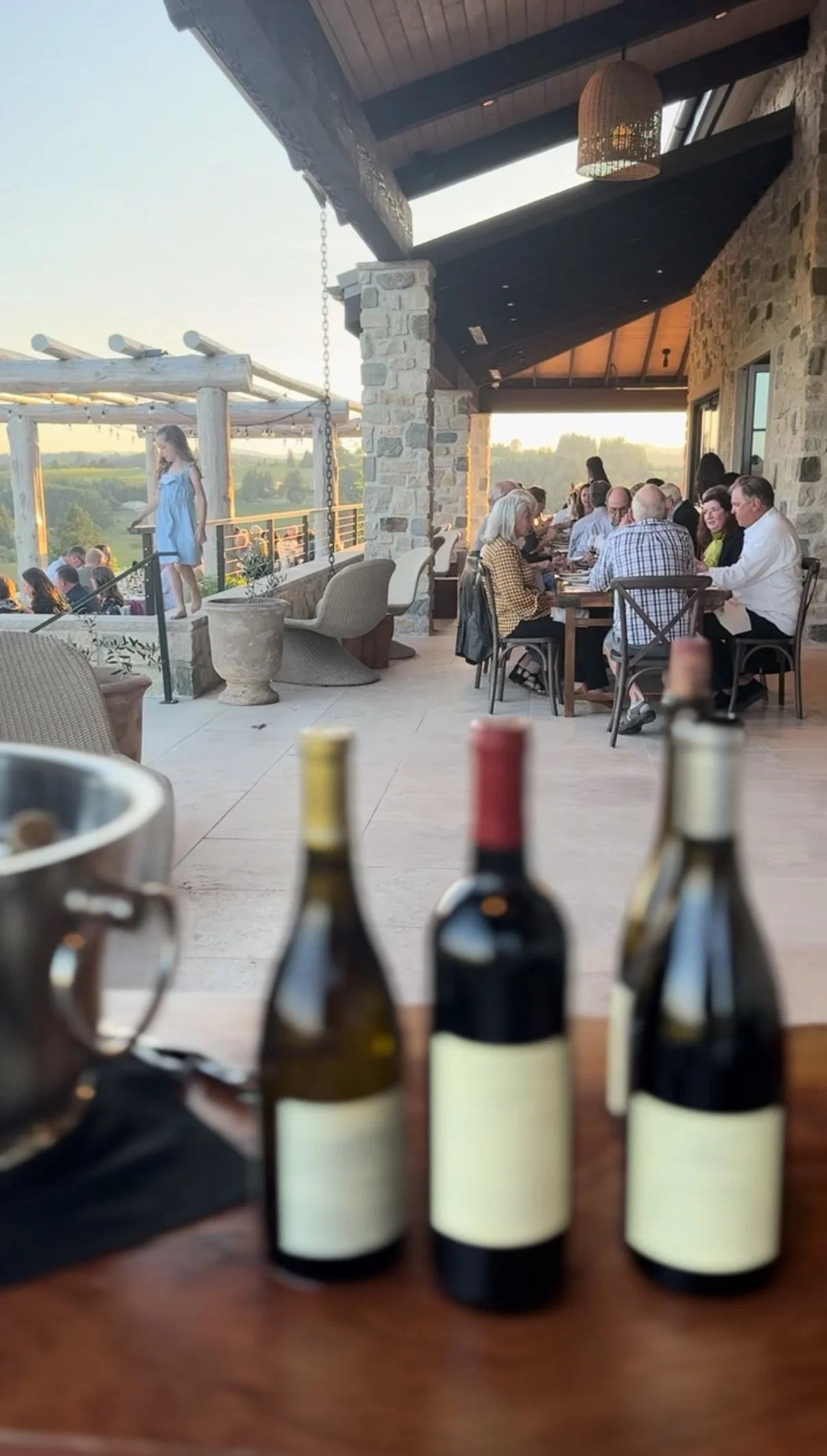 People dining on a terrace with stone pillars, a wooden ceiling, and a scenic view at sunset; three bottles of wine on the table in the foreground.