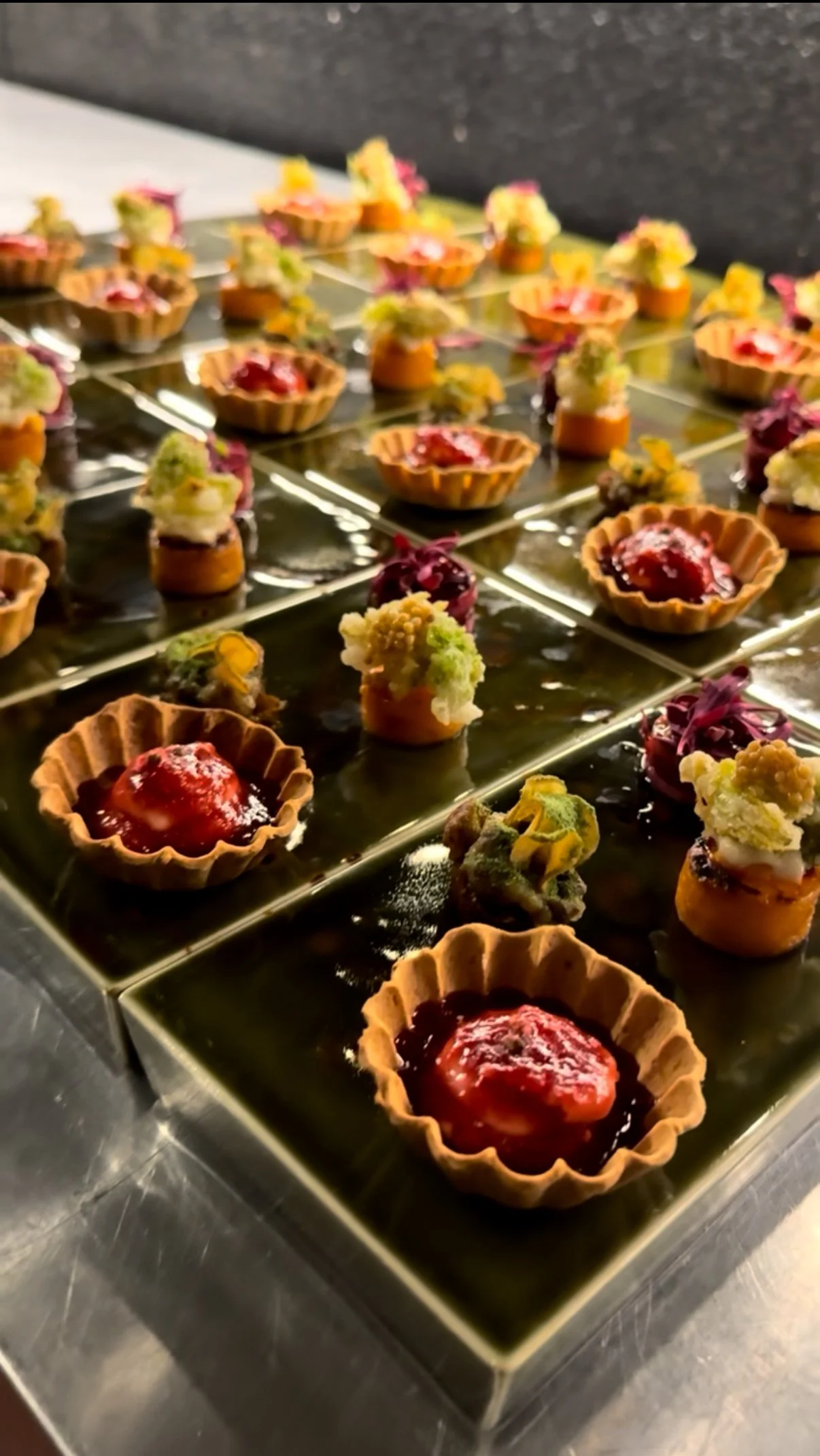 Close-up of assorted mini desserts on a reflective metallic serving tray, including tart shells with red fruit filling and various colorful, intricately decorated bite-sized pastries.