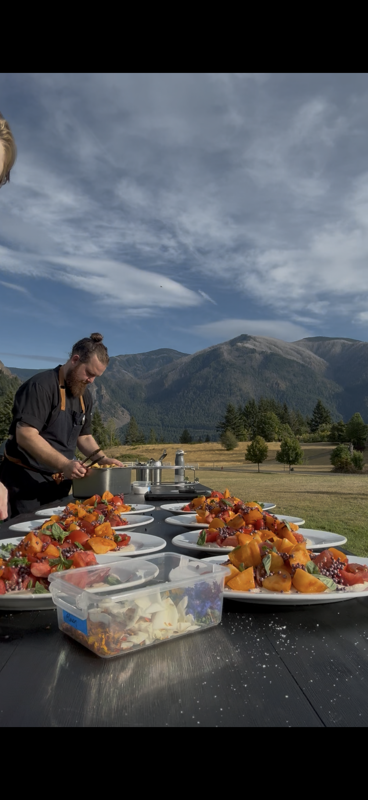 Chef preparing multiple plates of fruit salad outdoors with mountains and trees in the background.