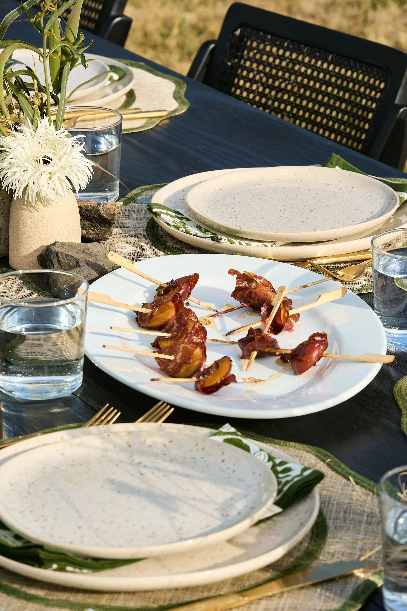 Outdoor table setting with plates, glasses of water, and a plate of bacon-wrapped dates with toothpicks, decorated with flowers, rocks, and green napkins.