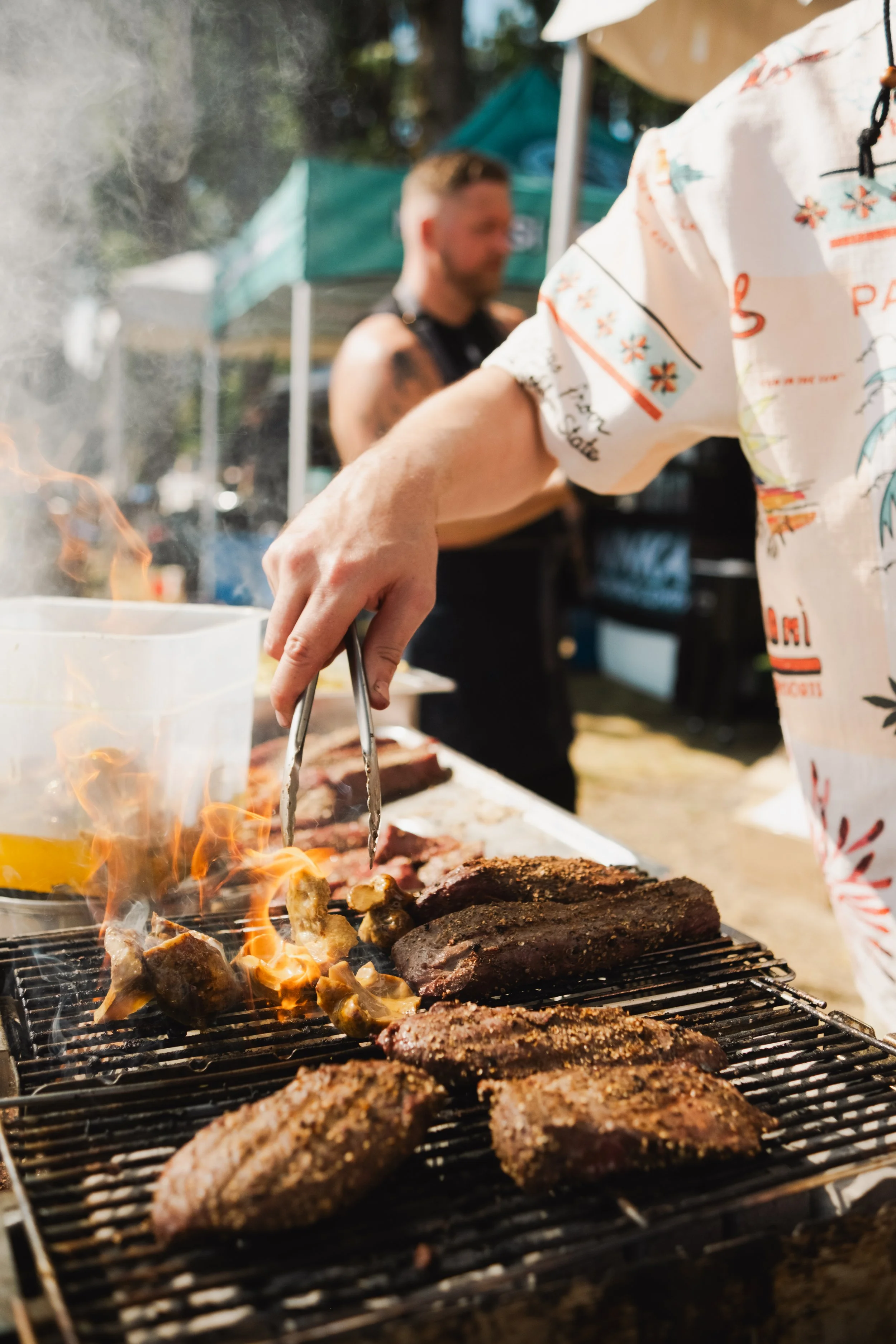 Person grilling various meats on a barbecue grill at an outdoor event, with another person in the background and tents overhead.