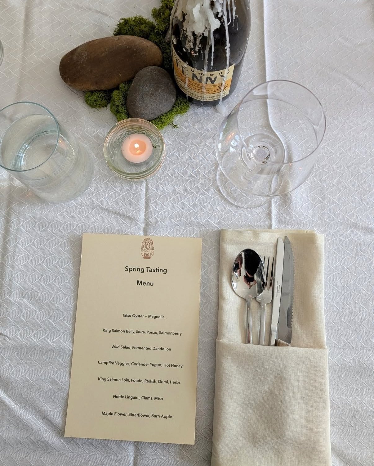 Table setting with a spring tasting menu, wine glass, water glass, cutlery wrapped in a napkin, and a candle, along with a decorative arrangement of rocks and moss with a partially extinguished bottle of wine or champagne.