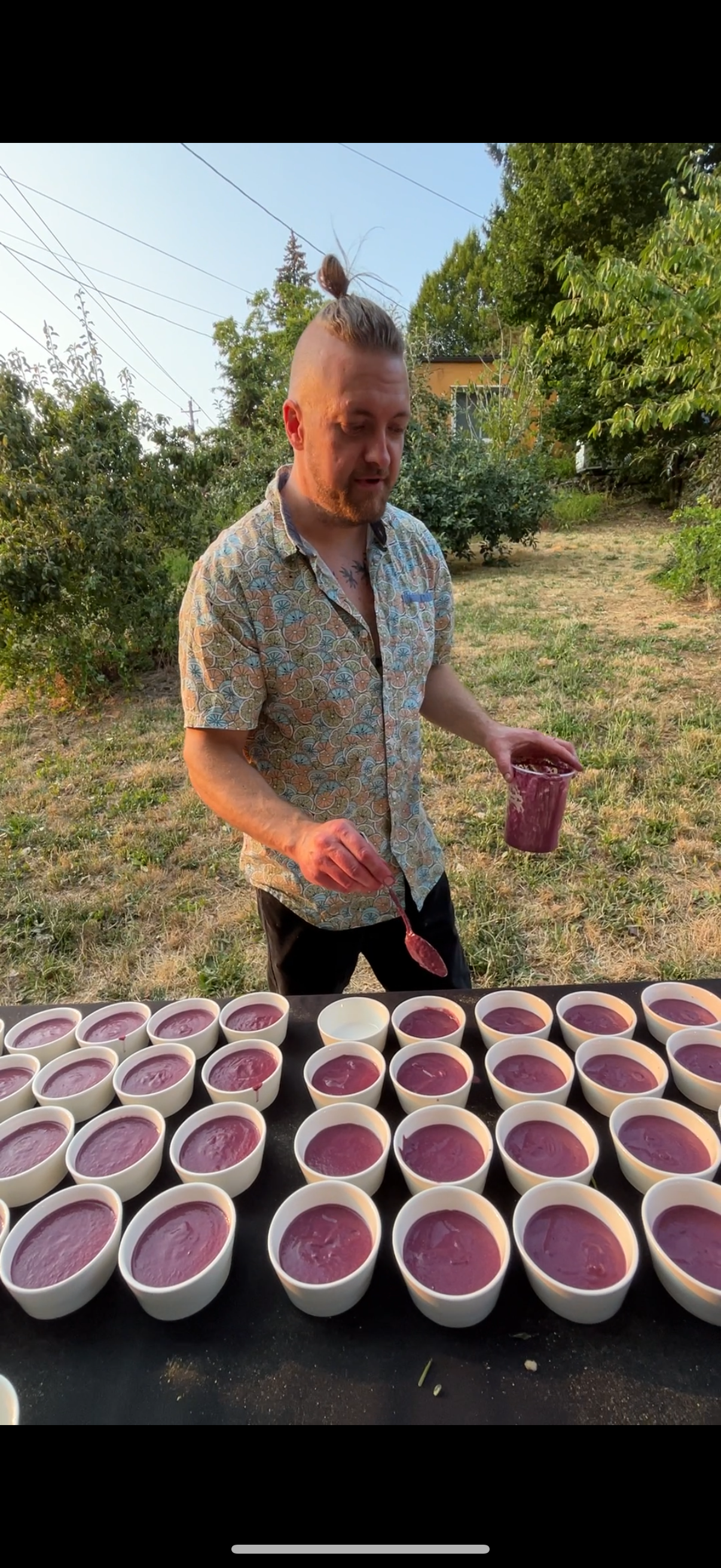 A man with a top knot hairstyle and a patterned short-sleeve shirt is serving a purple-colored dessert into small bowls on a black table outdoors, with greenery and trees in the background.