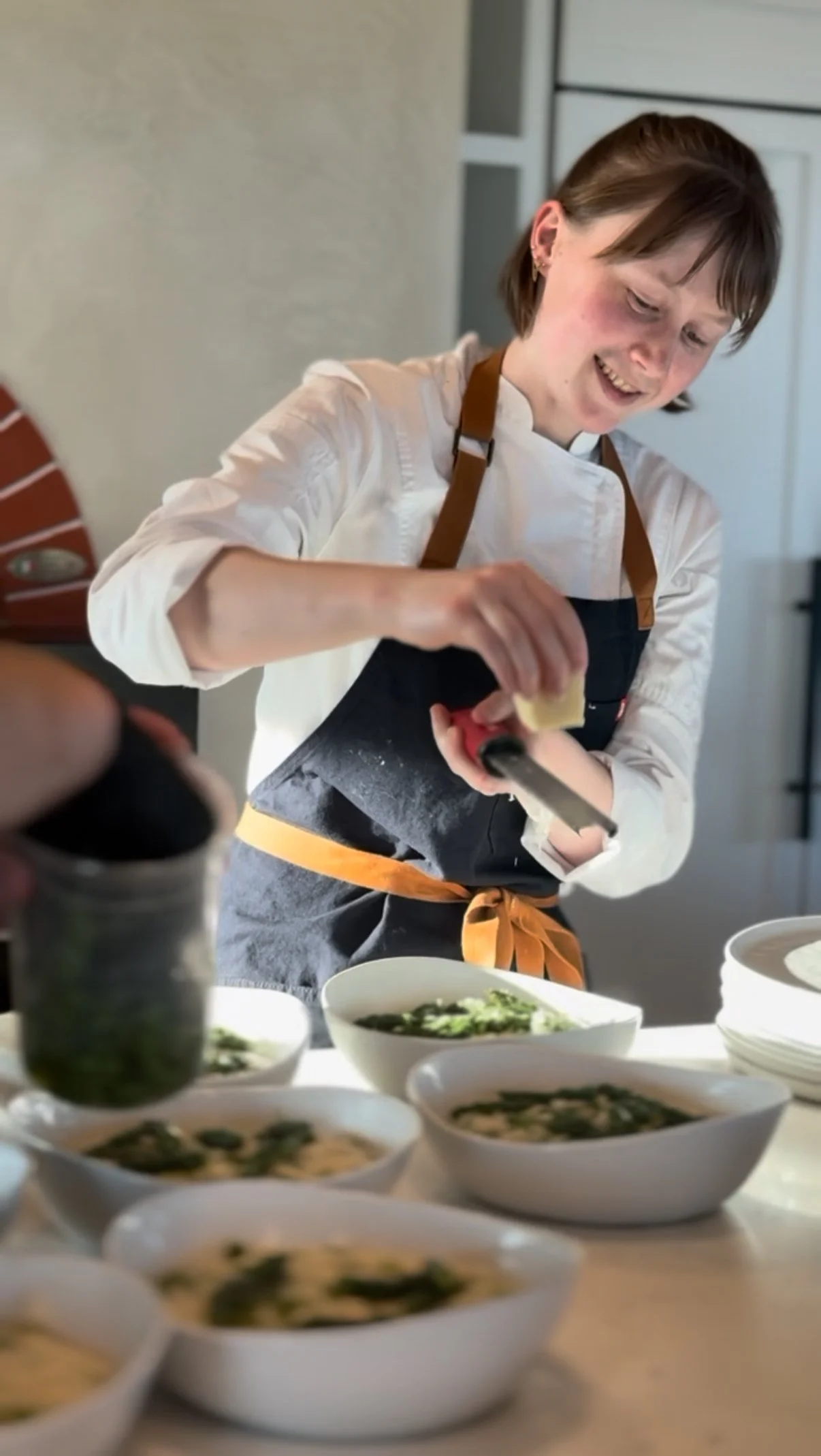 A woman in a white chef's coat and black apron is garnishing bowls of soup with herbs in a kitchen.