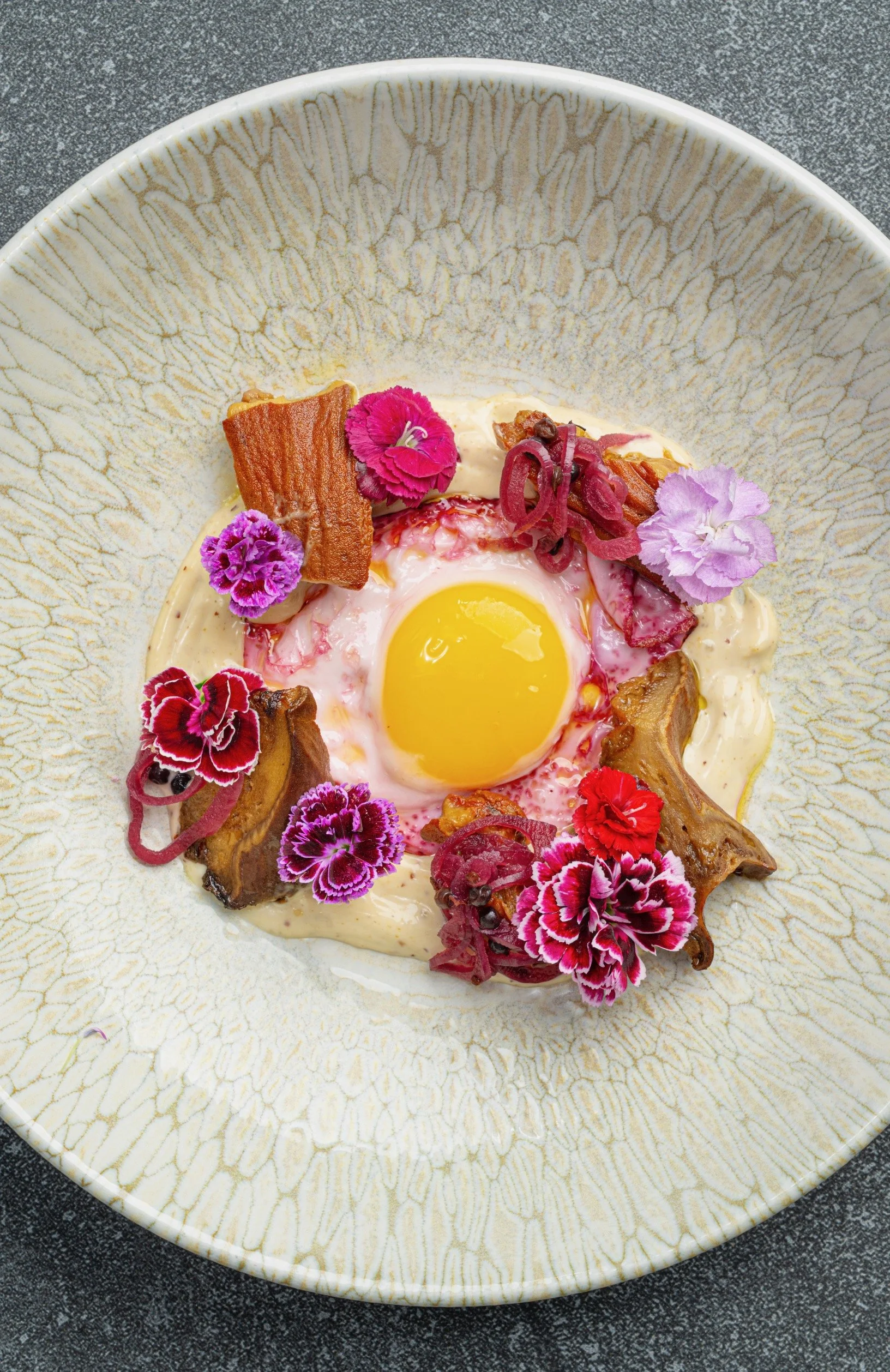 A bowl with a raw egg in the center, surrounded by colorful edible flowers, bacon, and caramelized pork, on a textured gray surface.