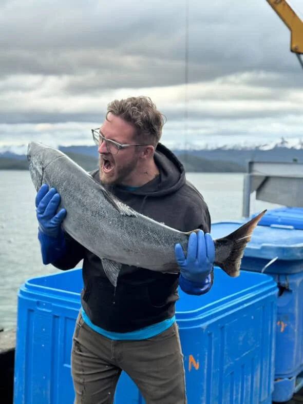 Man wearing glasses, a gray hoodie, and blue gloves, holding and pretending to bite a large fish outdoors near a body of water with mountains and cloudy sky in the background.