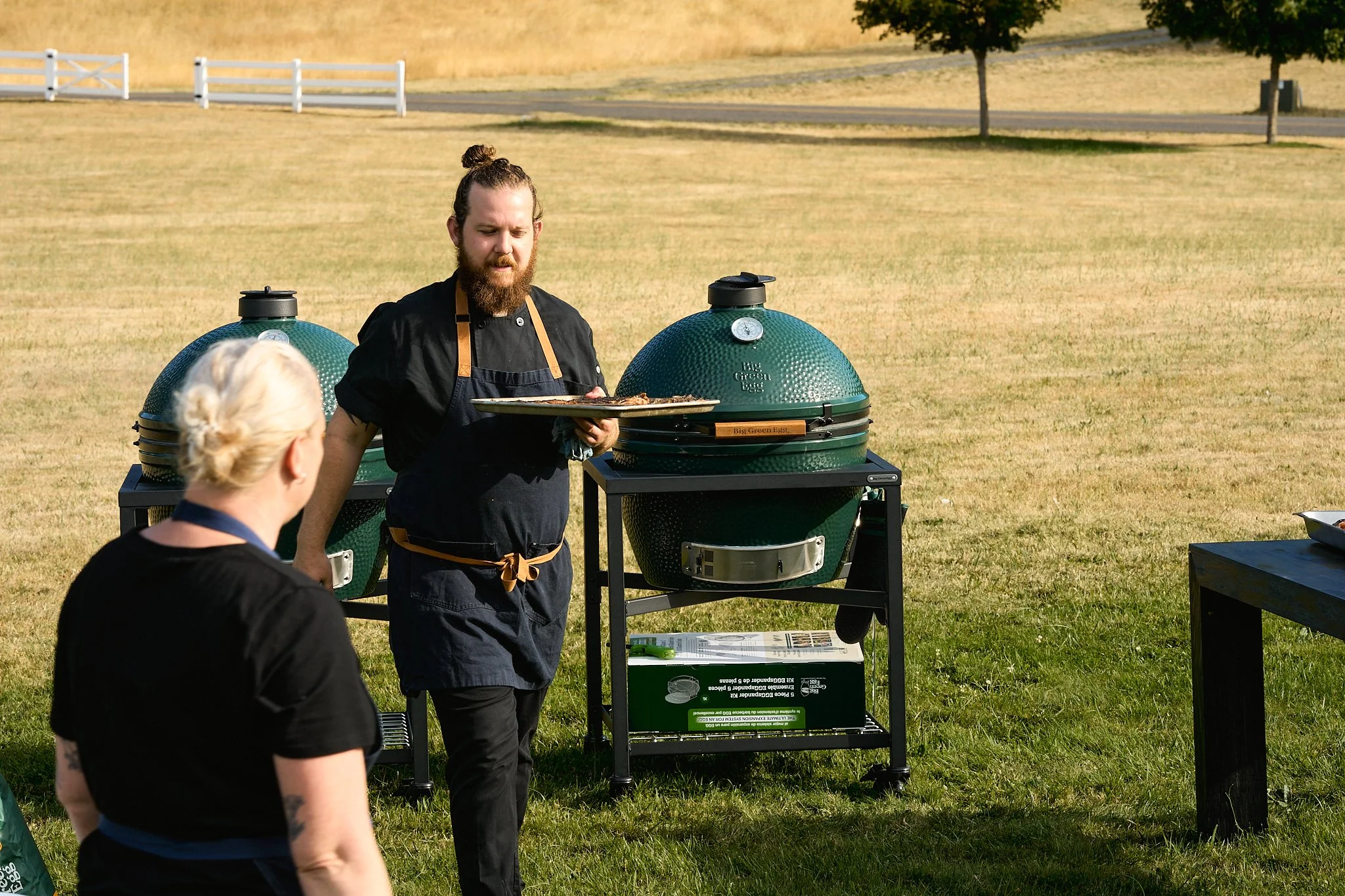 A man with a beard and long hair tied in a bun, wearing a black chef coat and apron, holds a tray of cooked food in front of two green outdoor smokers in a grassy field. A woman with short blonde hair wearing a black shirt is seated facing him, with 