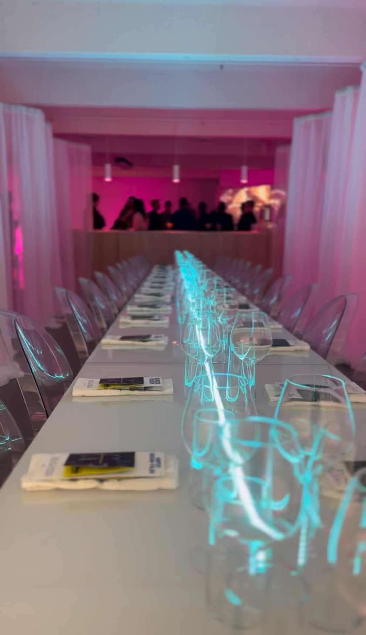 A long conference table set with wine glasses, plates, and napkins in a modern event space with pink and purple lighting and people gathered in the background.